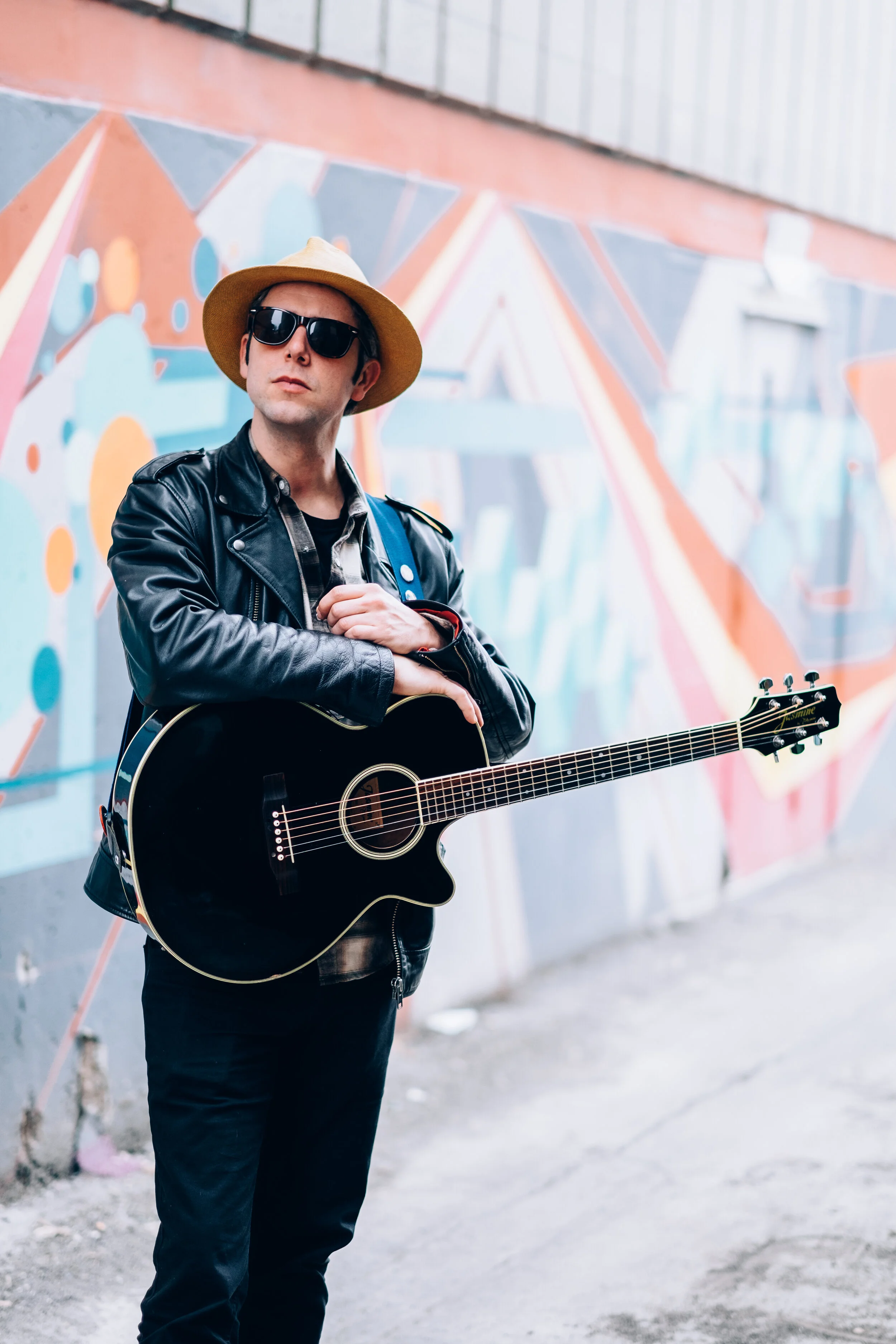 A young man wearing black sunglasses, a tan hat, and a black leather jacket, holding an acoustic guitar, standing in front of a colorful graffiti wall.