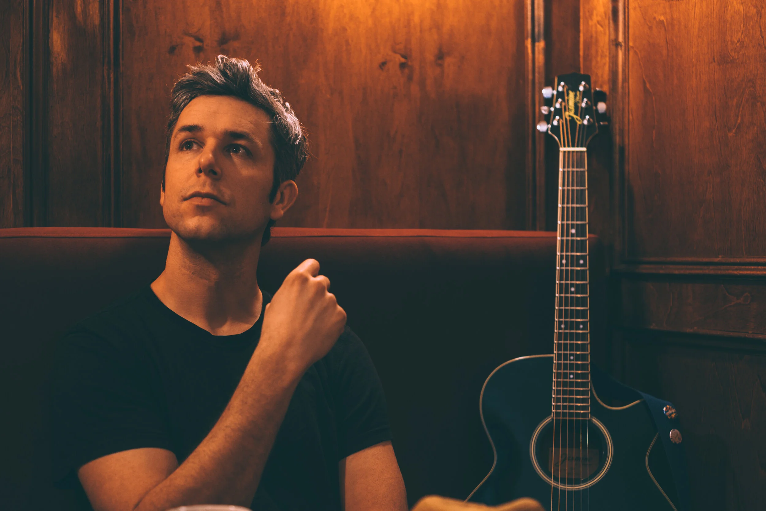 A man sitting in a dimly lit room with wooden walls, looking thoughtful. A guitar leans against the wall beside him.