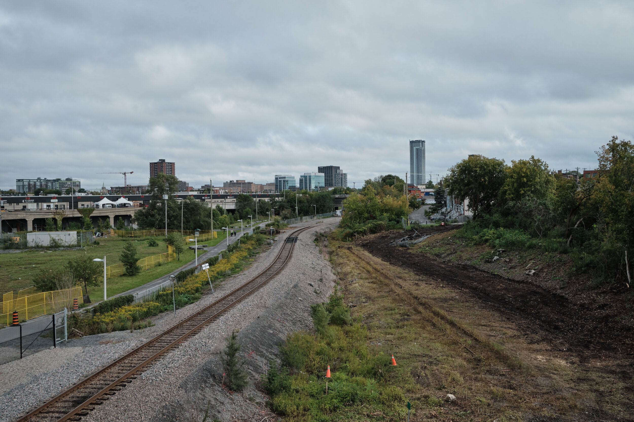 A cityscape view with train tracks curving through an area under development, with trees, construction cones, and distant buildings under a cloudy sky.