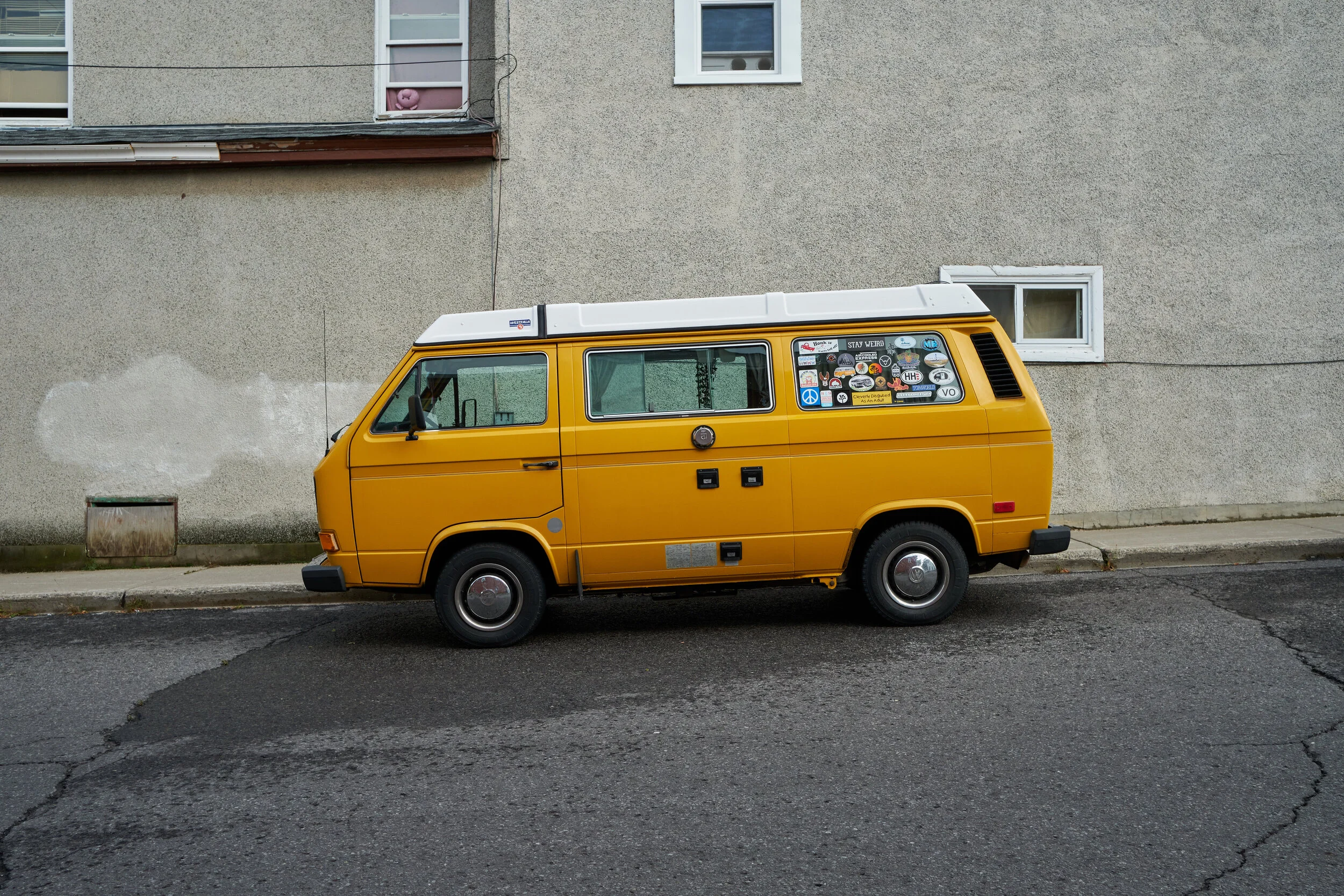 A yellow vintage camper van parked on the street in front of a beige building with two small windows, one of which has pink toys inside, and one larger window with stickers on the outside.