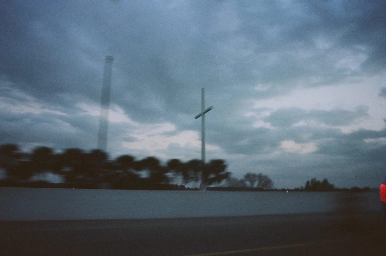 Overcast sky with dark gray clouds over a highway, a utility pole with a crossbeam, and trees in the background. Blurred motion suggests the photo was taken from a moving vehicle.