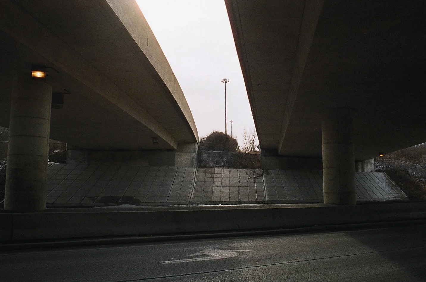 Underpass with concrete support columns and a bridge overhead, seen from a vehicle on the road underneath on a cloudy day.
