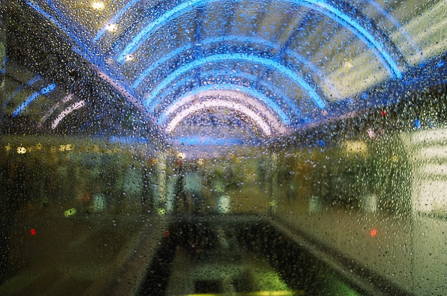 Night view through a rain-covered window of an illuminated curved passage with blue and purple lights and green and yellow lights in the background.