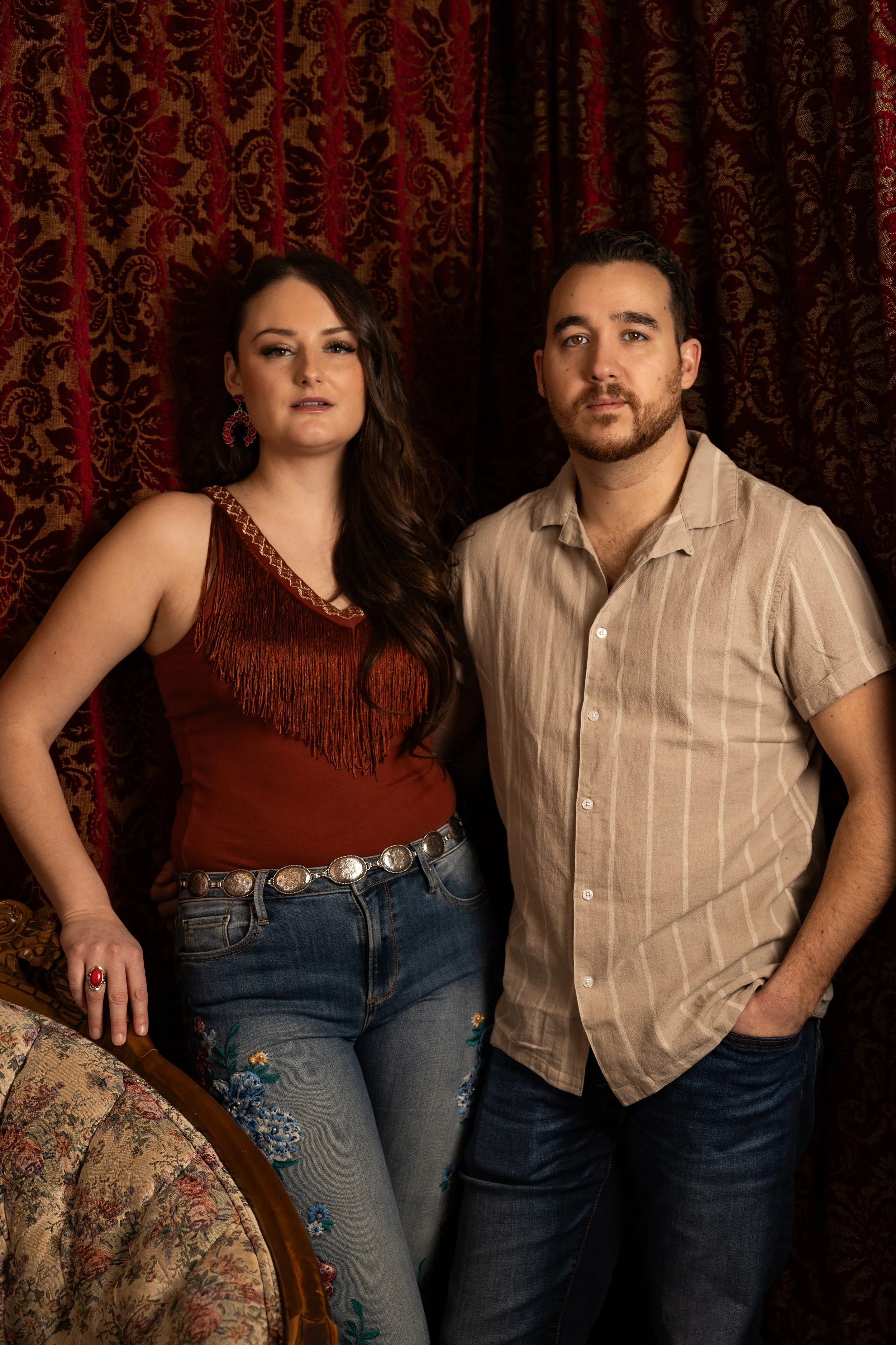 A man and woman standing in front of a red and gold patterned curtain, posing for a photo indoors.
