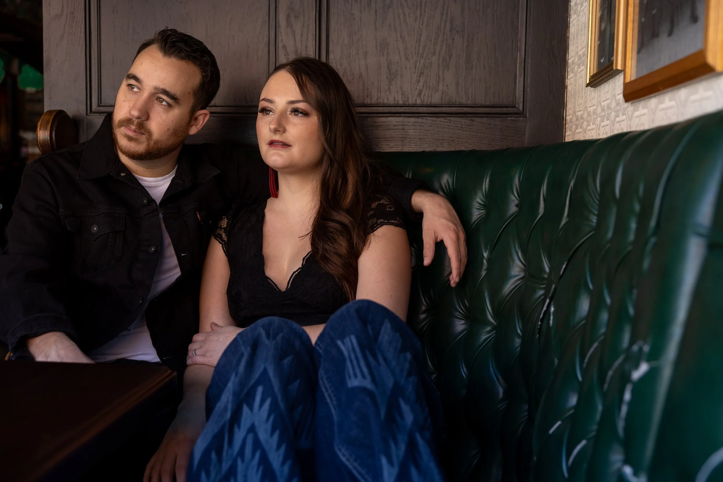 A man and woman sitting closely on a green leather booth in a dimly lit restaurant or bar, looking in opposite directions with serious expressions.