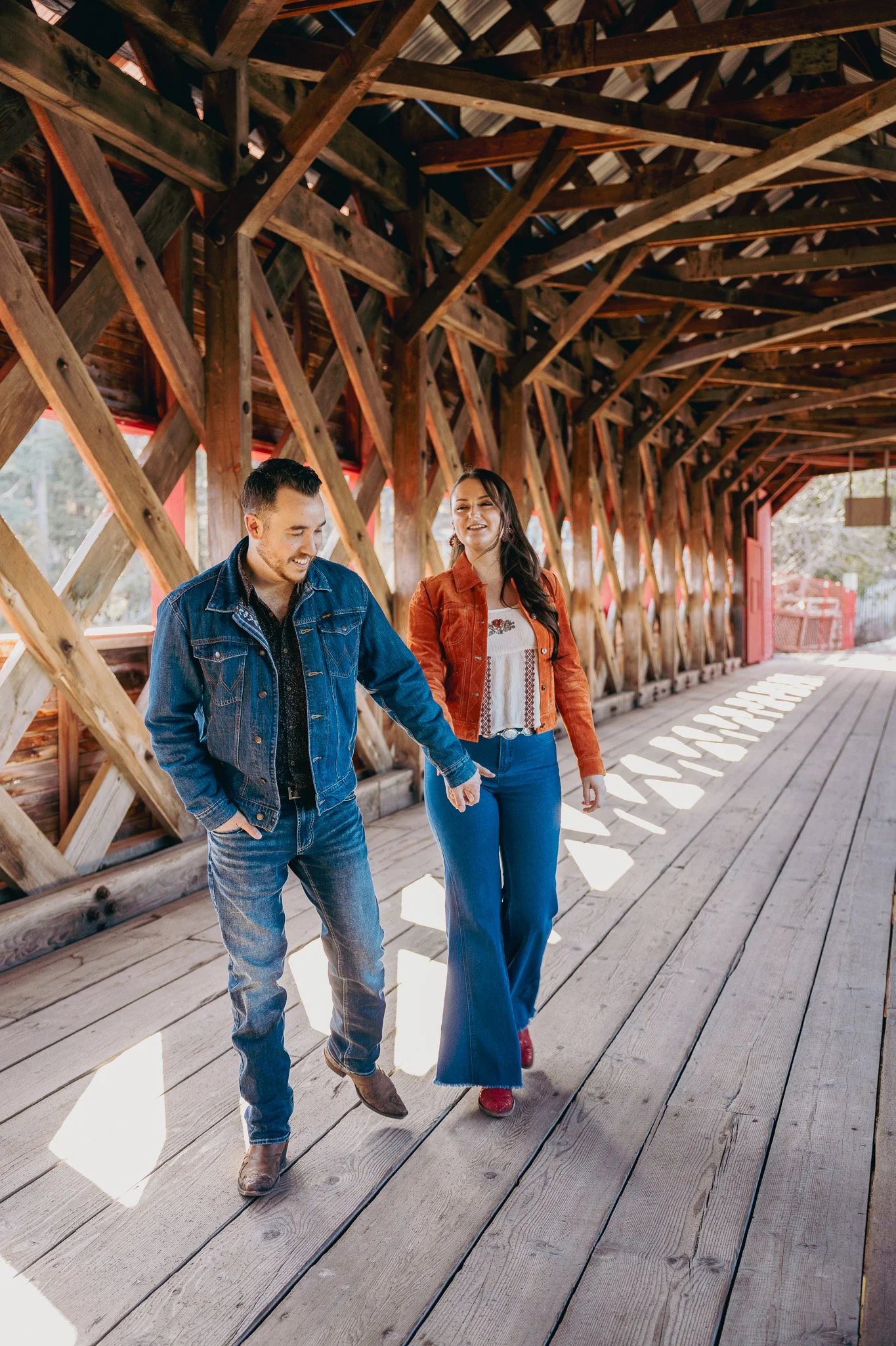 A man and woman holding hands walking on a wooden covered bridge with sunlight streaming through gaps in the side walls.