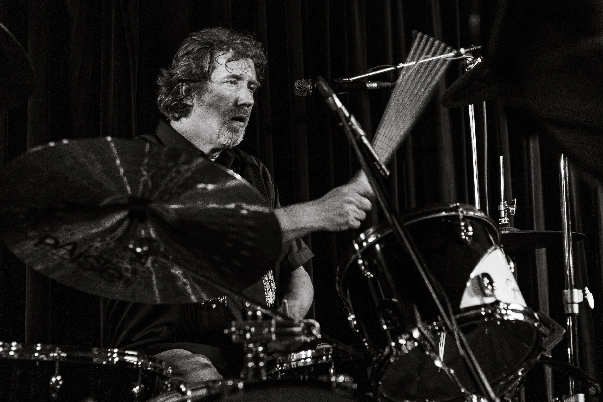 Black and white photo of a man playing drums on stage with a black curtain behind him.