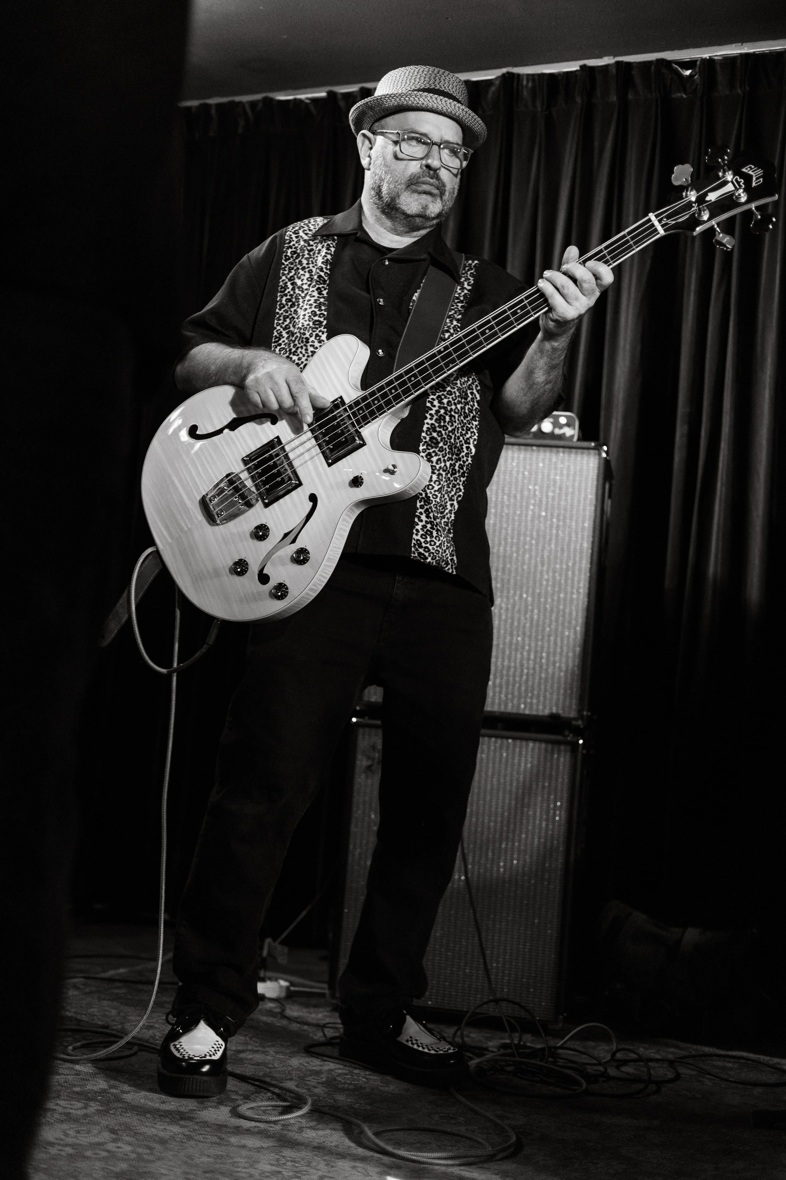 Black and white photo of a man playing an electric guitar on stage, wearing glasses, a hat, a black shirt with patterned accents, and standing in front of a dark curtain and amplifier.
