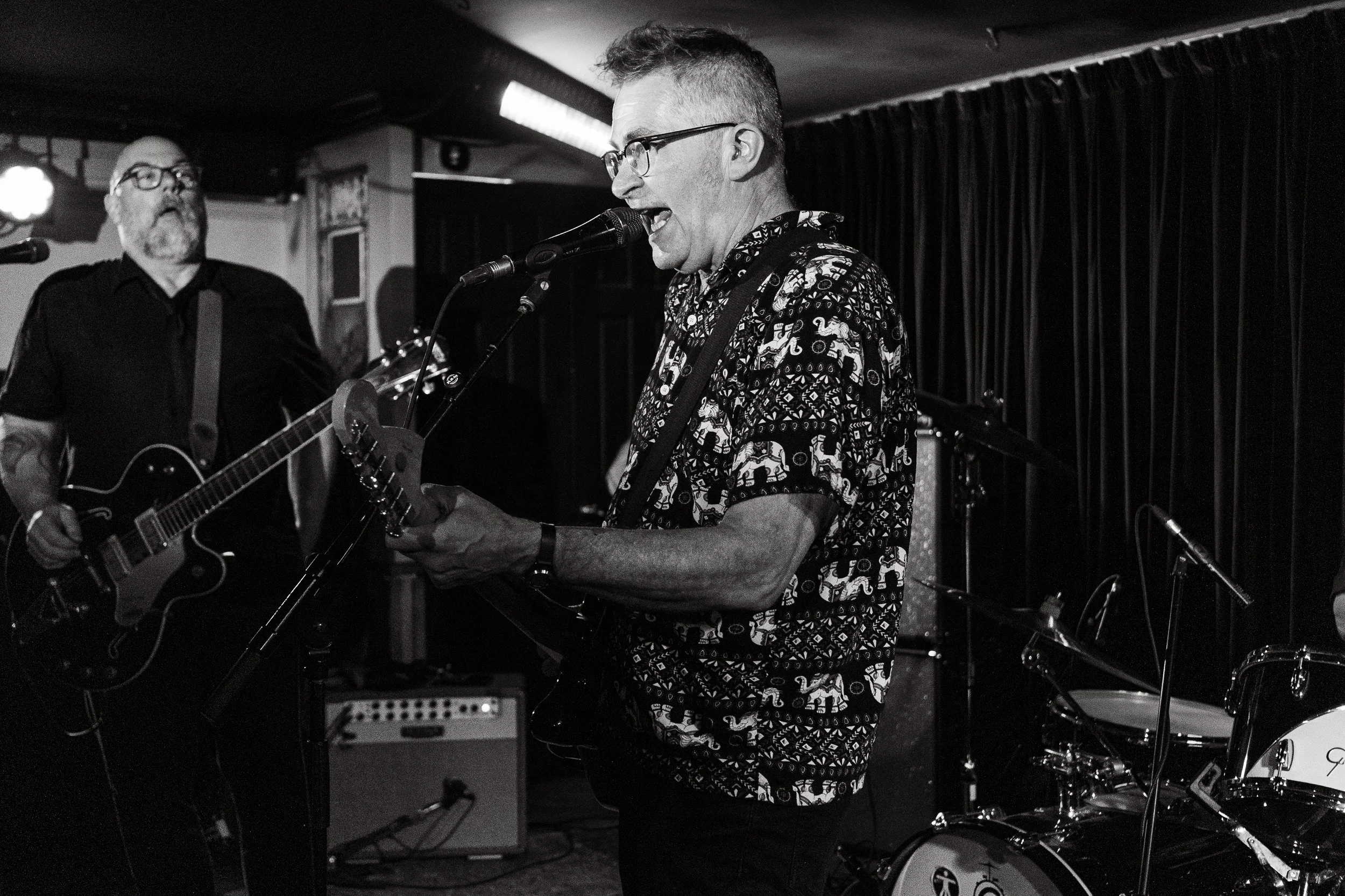 Black and white photo of two men performing on stage; one man is playing guitar and singing into a microphone, wearing a patterned shirt, glasses, and with short hair; the other man is playing guitar in the background, wearing glasses, a dark shirt, 