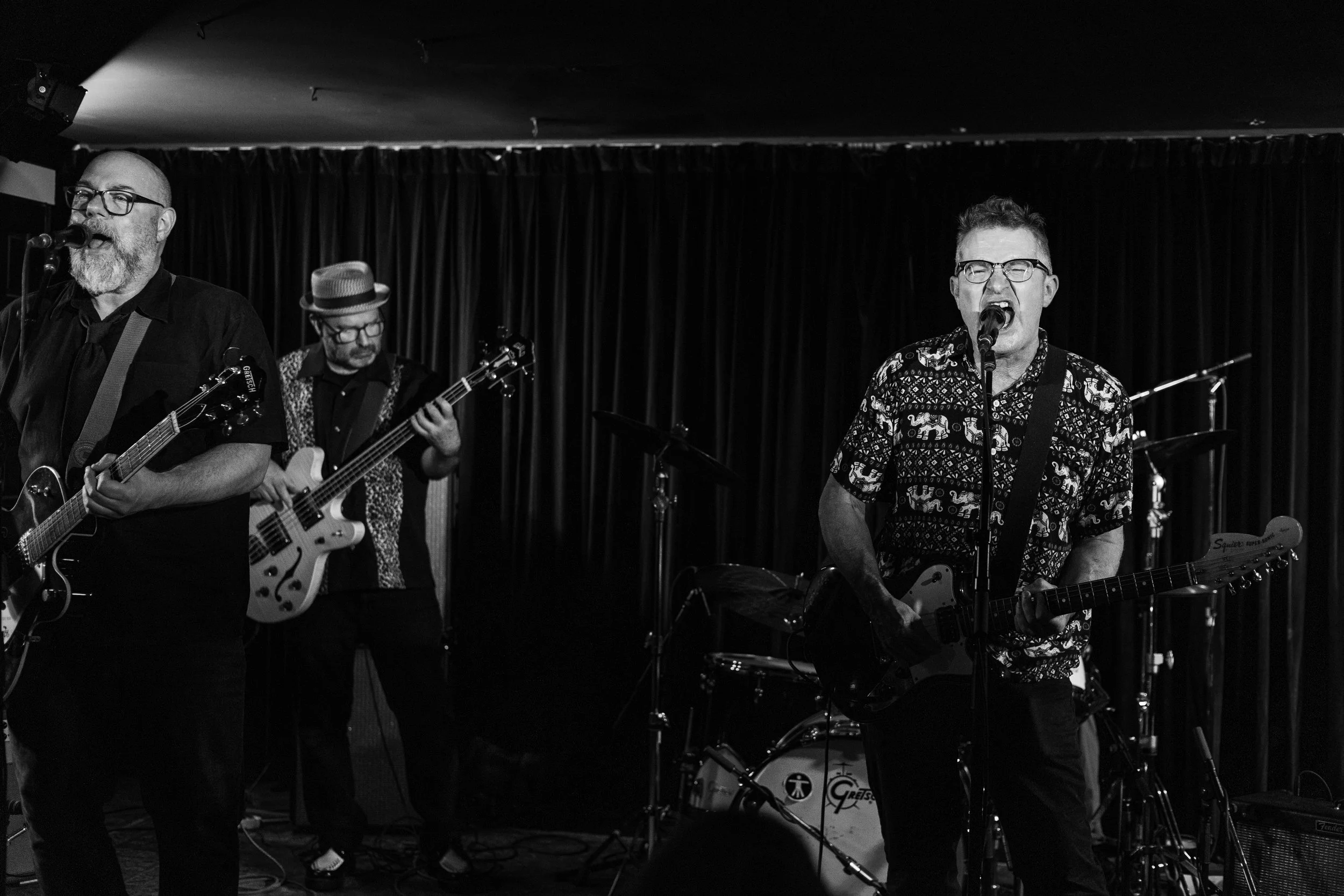 A black and white photo of a band performing on stage with three male musicians playing guitars and drums in the background.