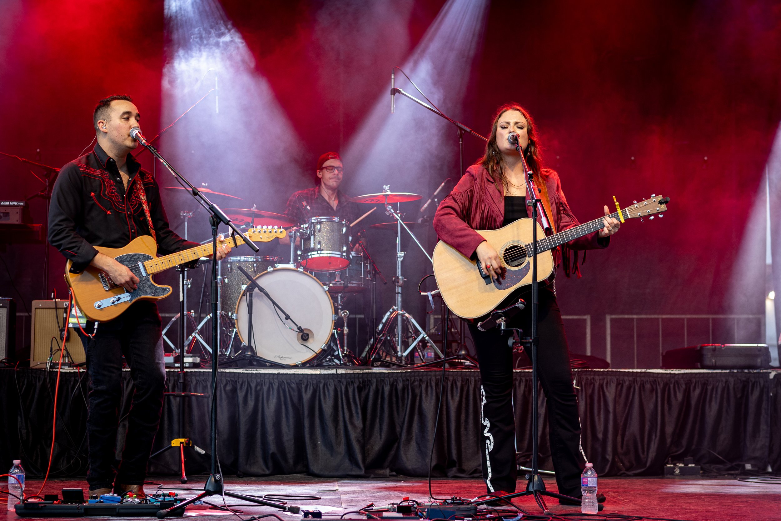 A band performs on stage with two guitarists and a drummer, under red and white stage lighting.