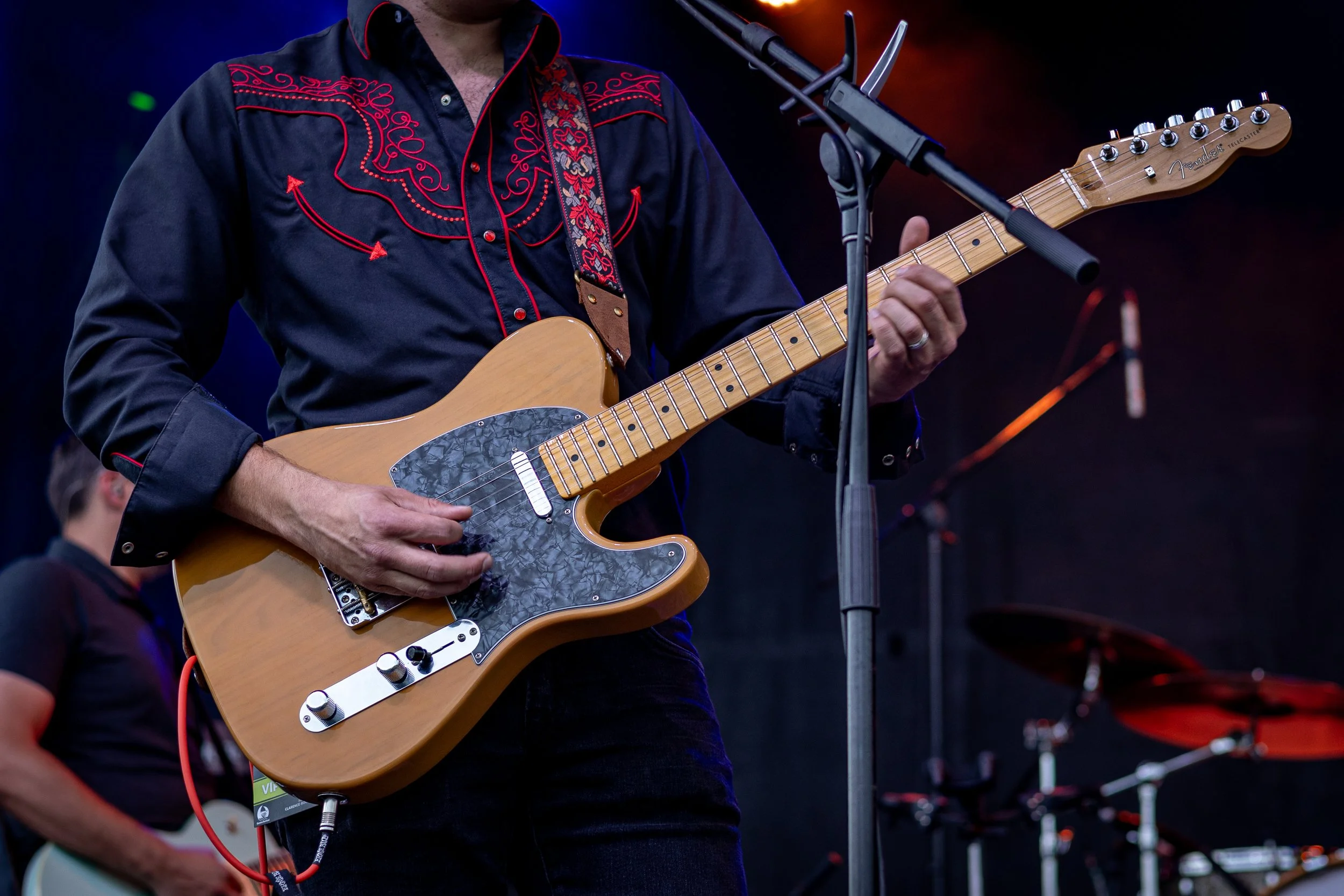 A musician playing an electric guitar on stage, dressed in a black Western-style shirt with red embroidery.