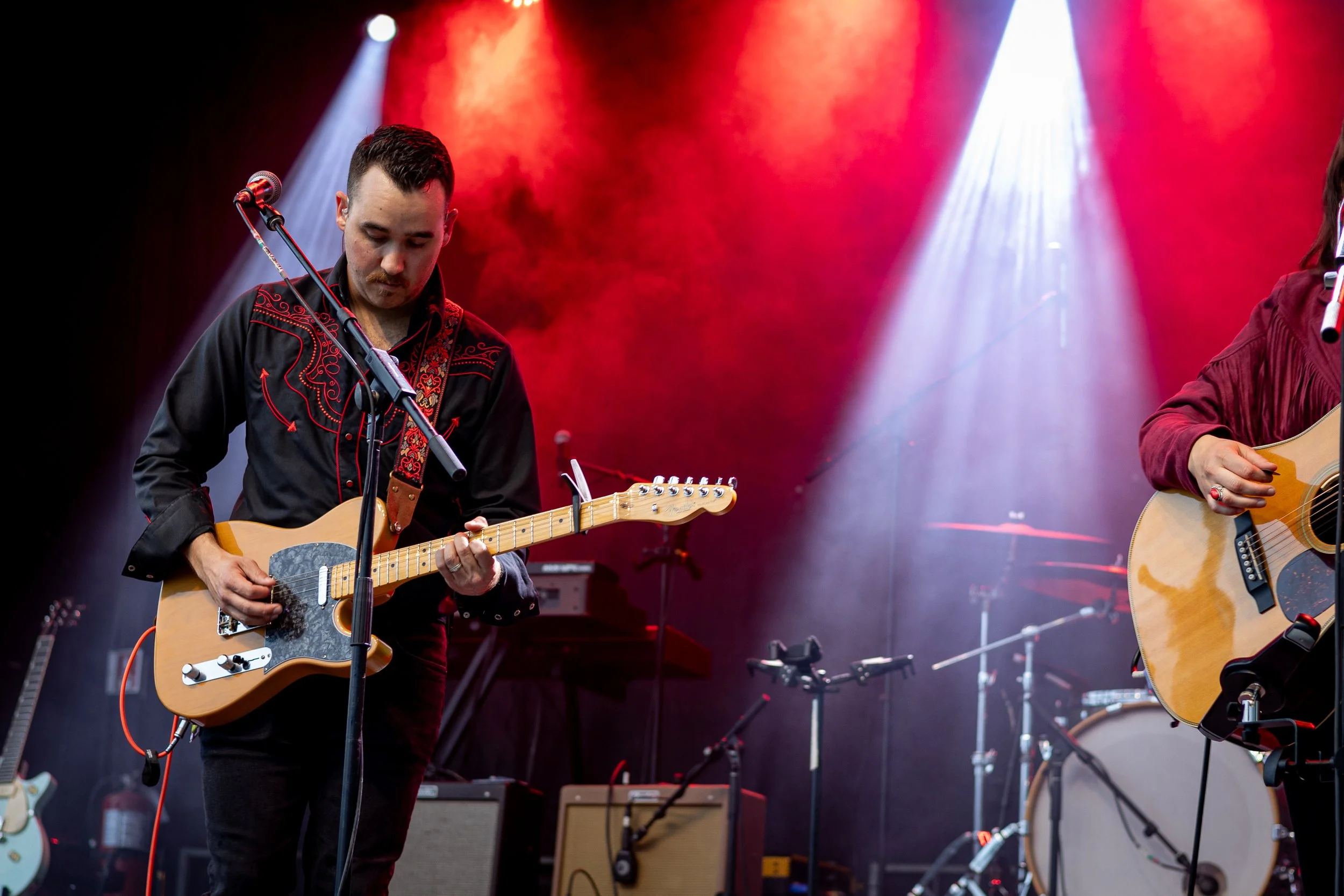 Two musicians playing guitars on stage with red and white stage lighting and musical equipment in the background.
