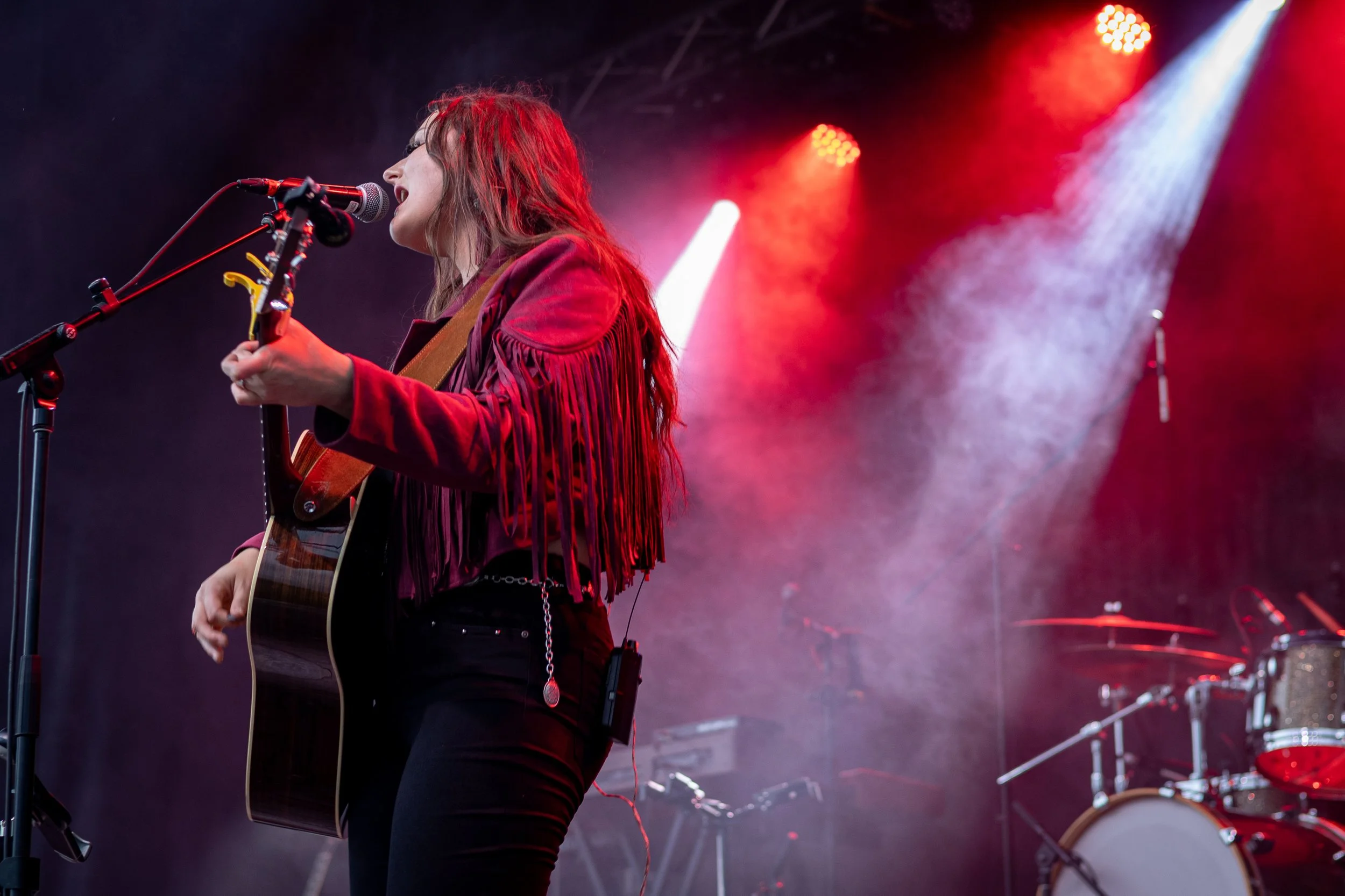 A female musician performing with a guitar on stage under red and white lights with a drum set in the background.