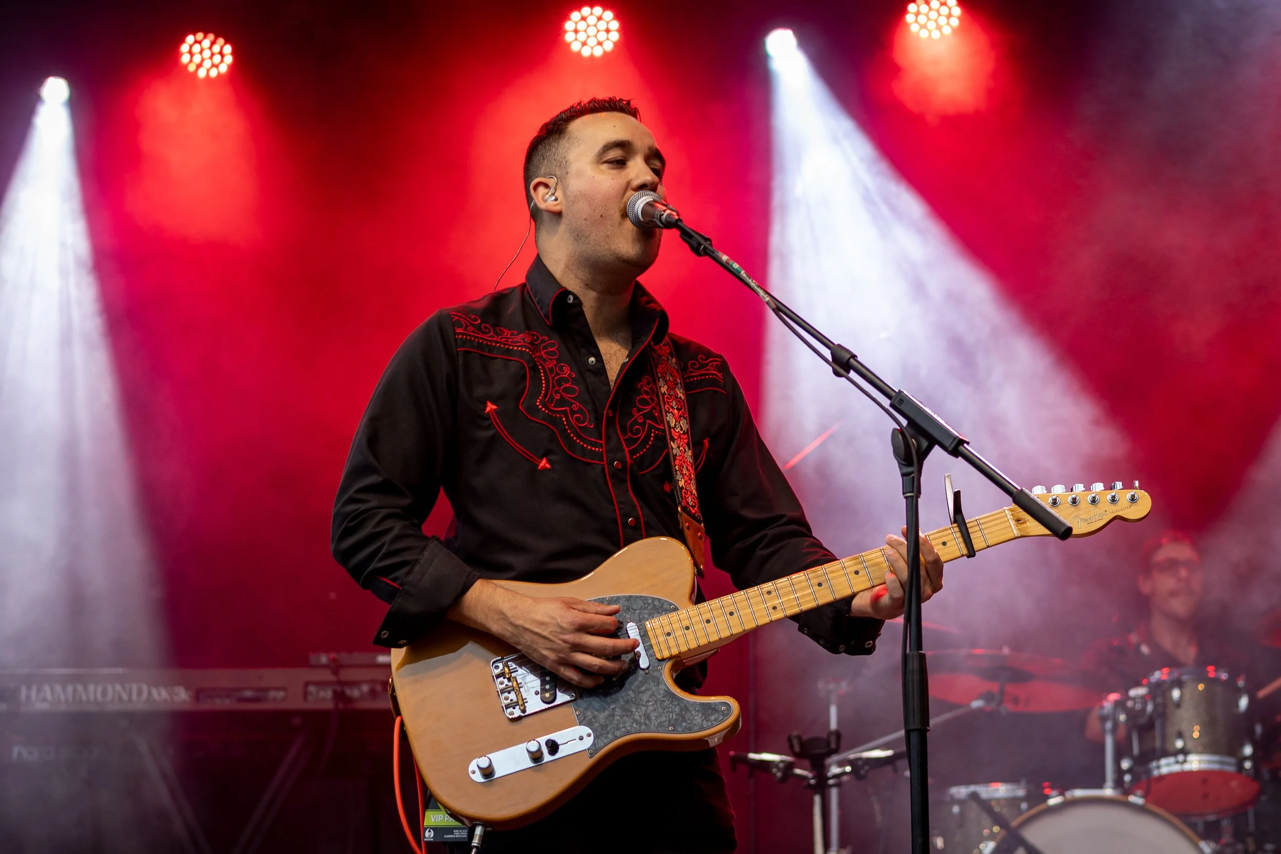 A musician performing on stage with a guitar, singing into a microphone, with red stage lights and a drummer in the background.