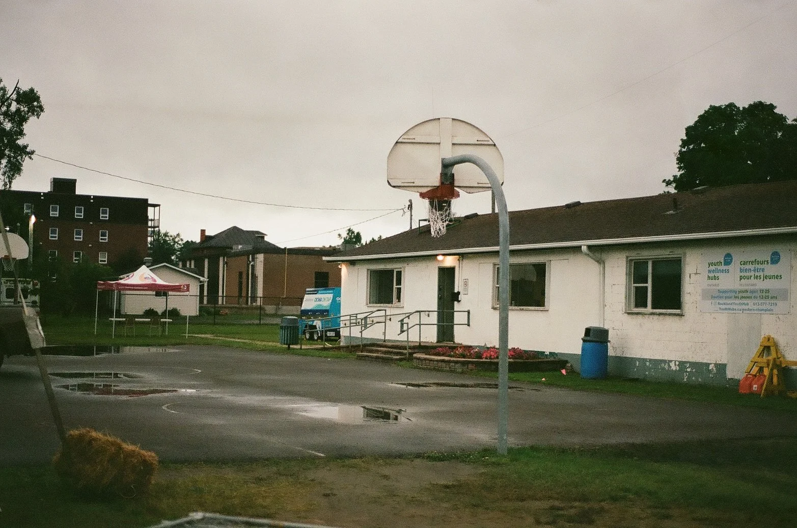 An outdoor basketball court with a backboard and hoop, wet pavement with puddles, a white building with a sign for youth wellness hubs, and tents in the background on a cloudy evening.