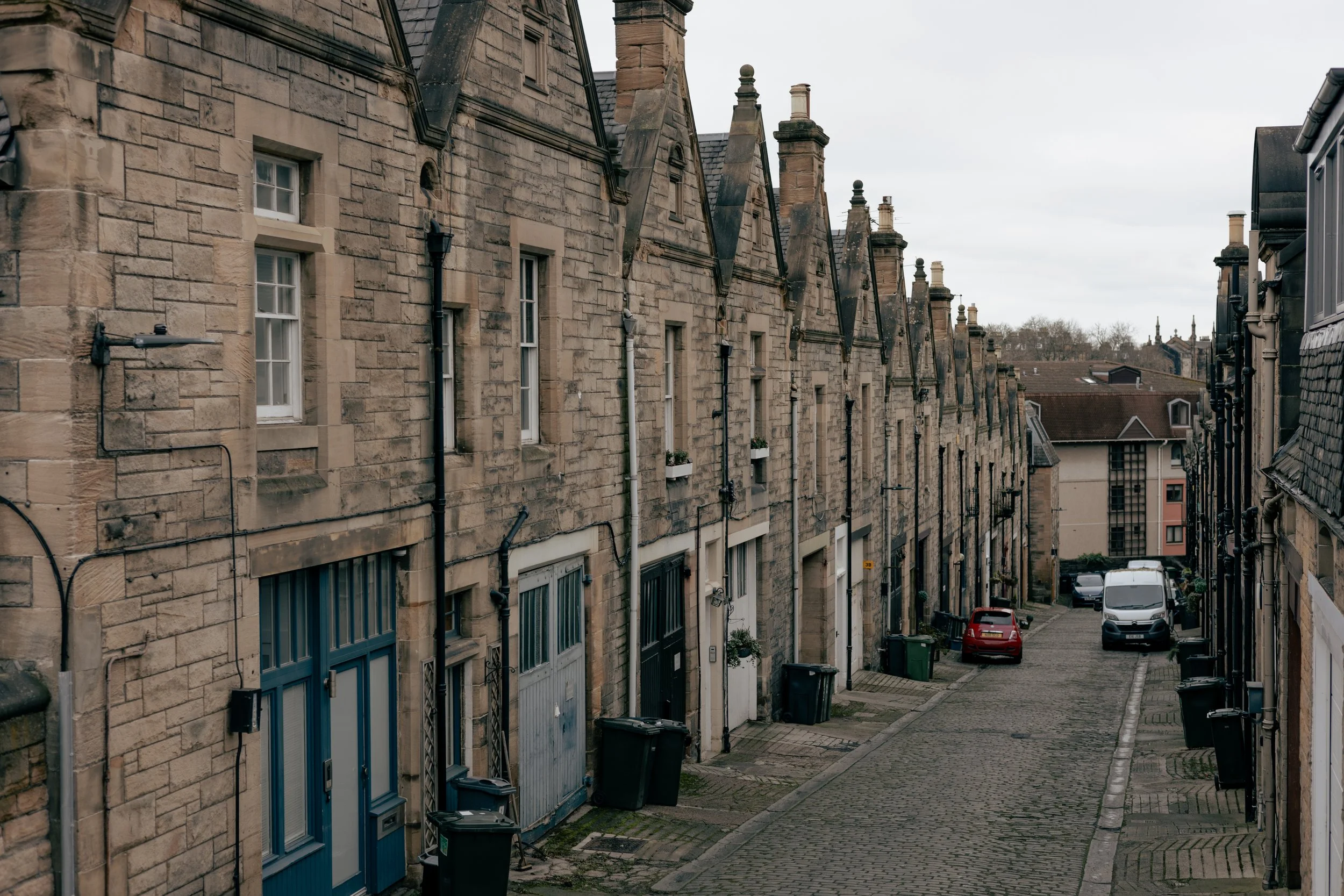 A narrow cobblestone street in a historic neighborhood with old stone row houses, some with small windows and flower boxes, parked cars, and trash bins along the sidewalk under an overcast sky.
