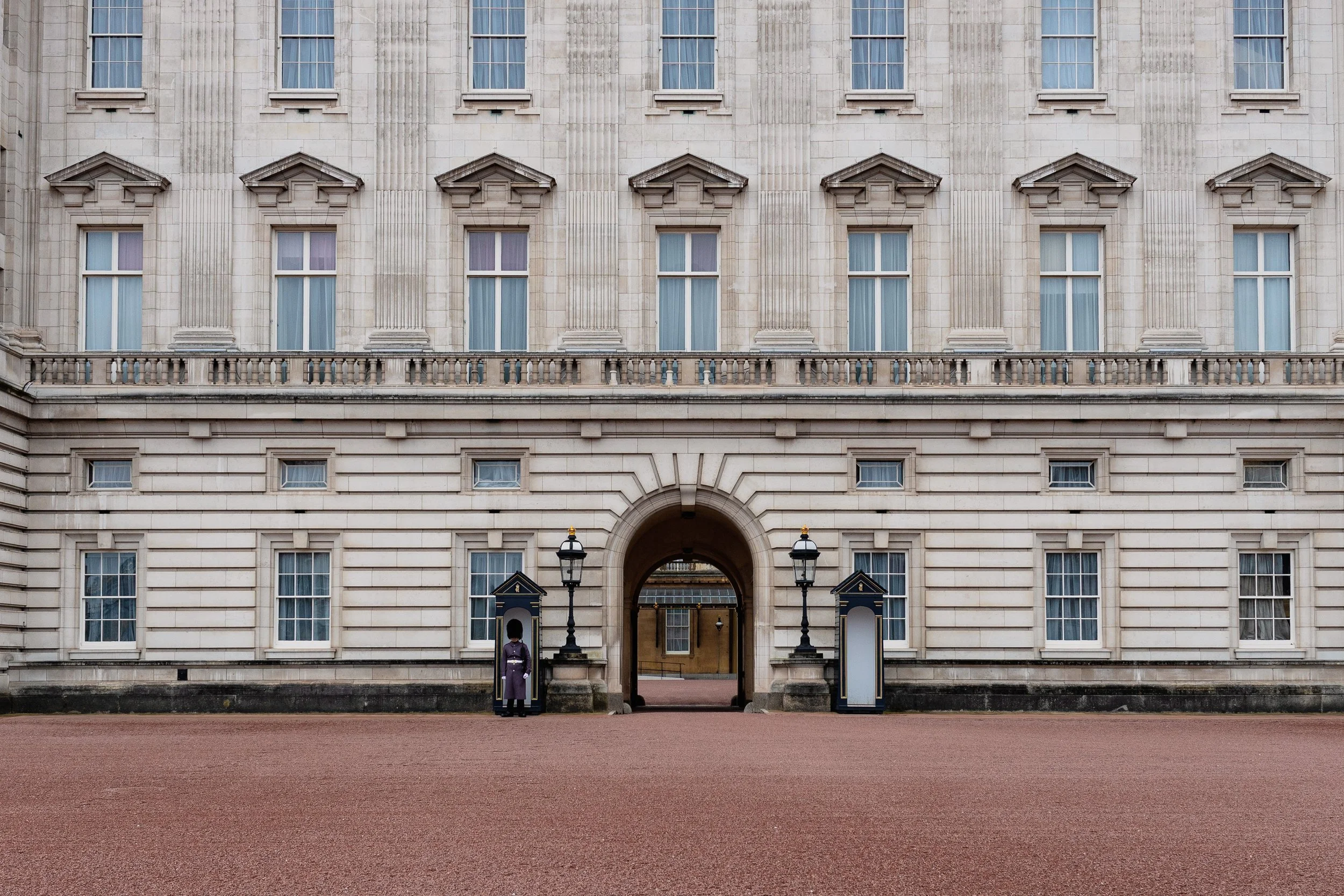 The facade of a large, historic, beige stone building with multiple windows, an arched entrance, and decorative architectural details. Two ceremonial guards stand on either side of the entrance, flanking guardhouses and lamps.