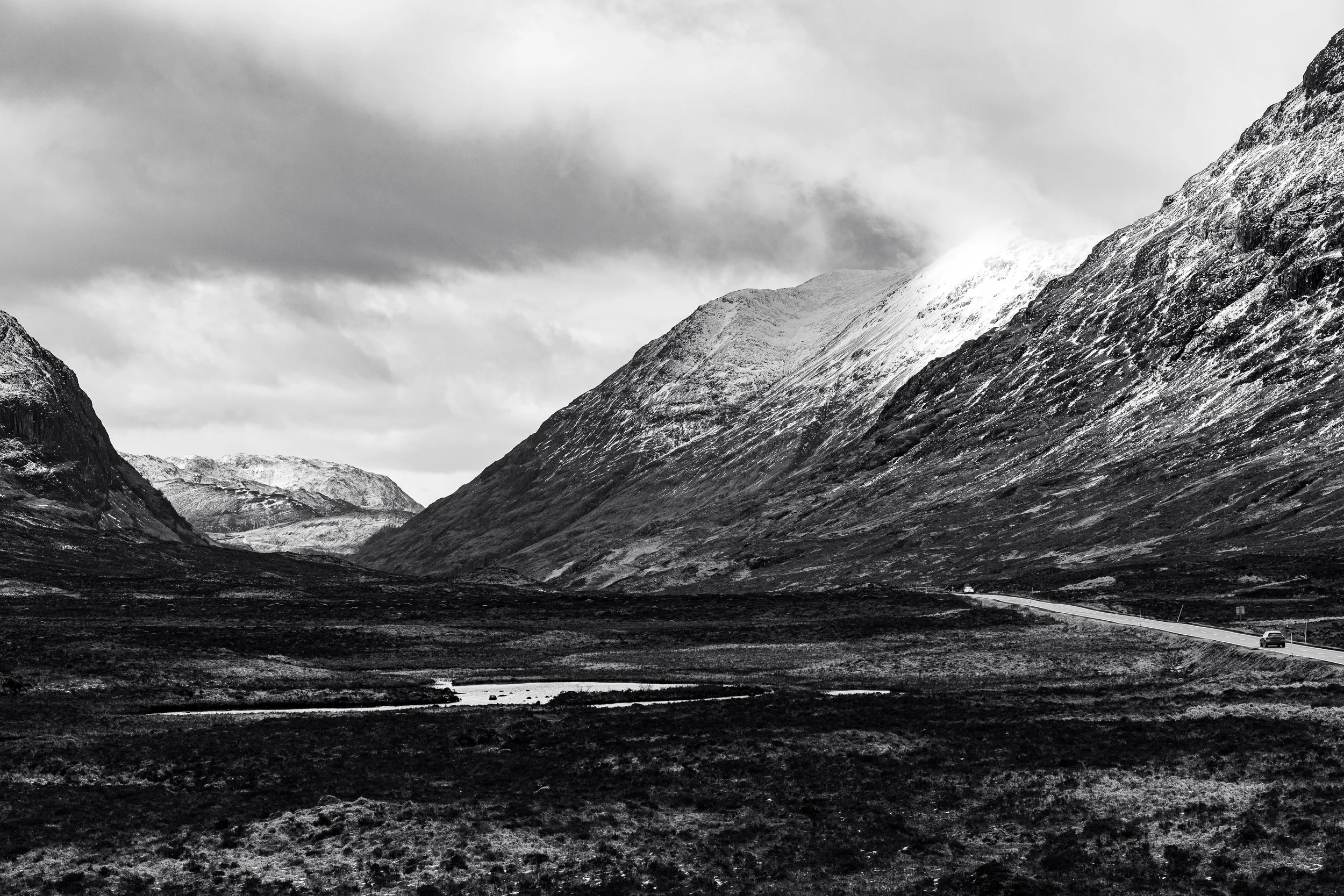 Black and white photograph of a mountainous landscape with snow-covered peaks, a winding road with cars, and a small body of water in the foreground.