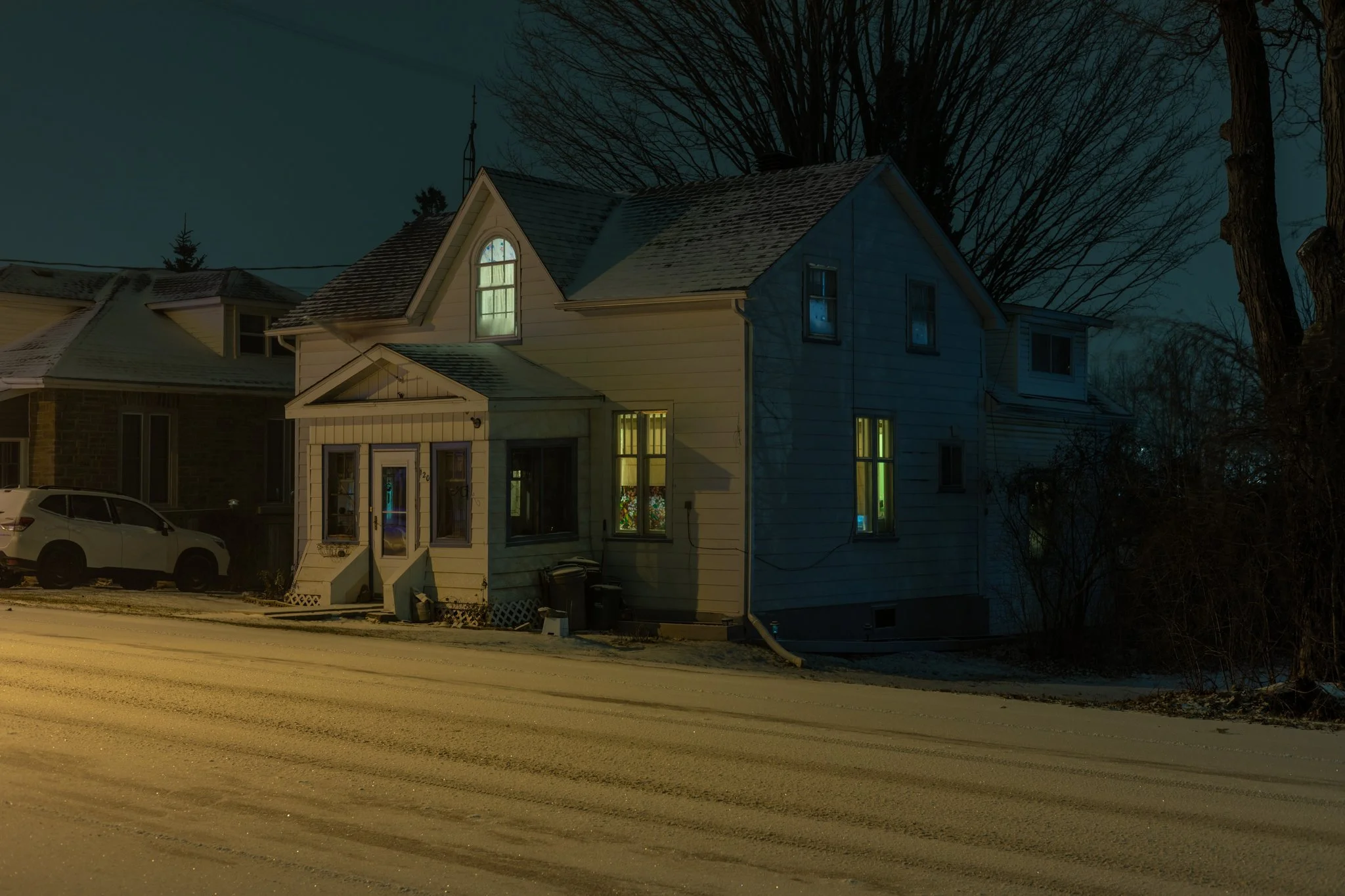 A house illuminated from within at night, with snow on the street and rooftops, and leafless trees in the background.