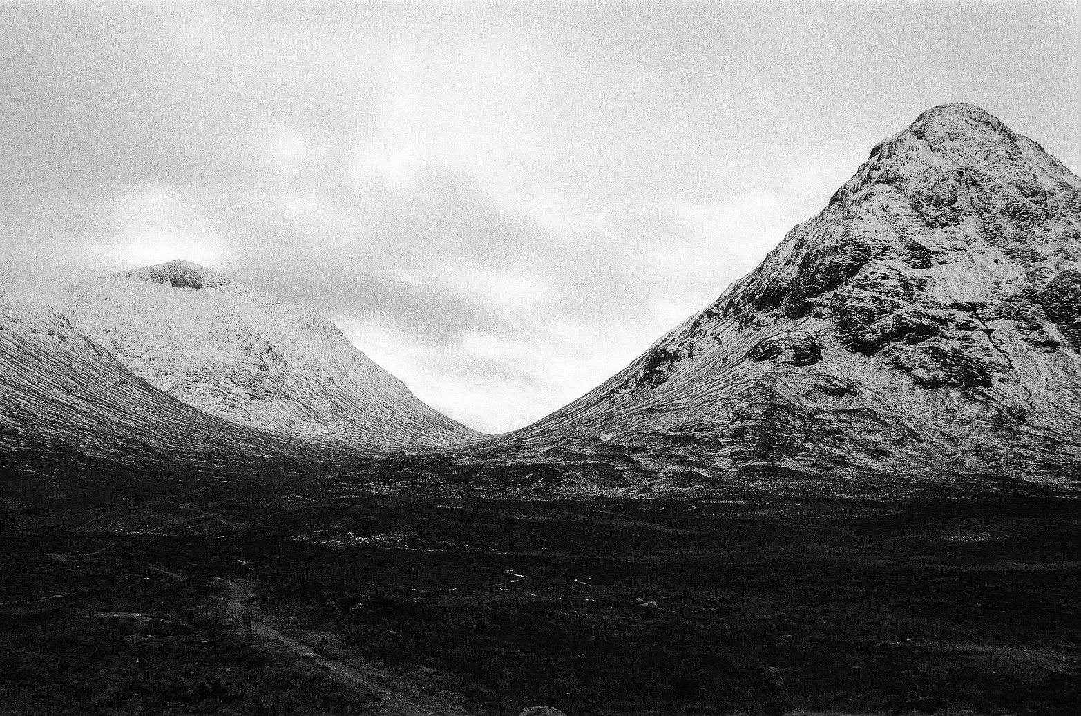 Black and white photograph of two snow-covered mountains with a cloudy sky above and a dark, rugged landscape below.