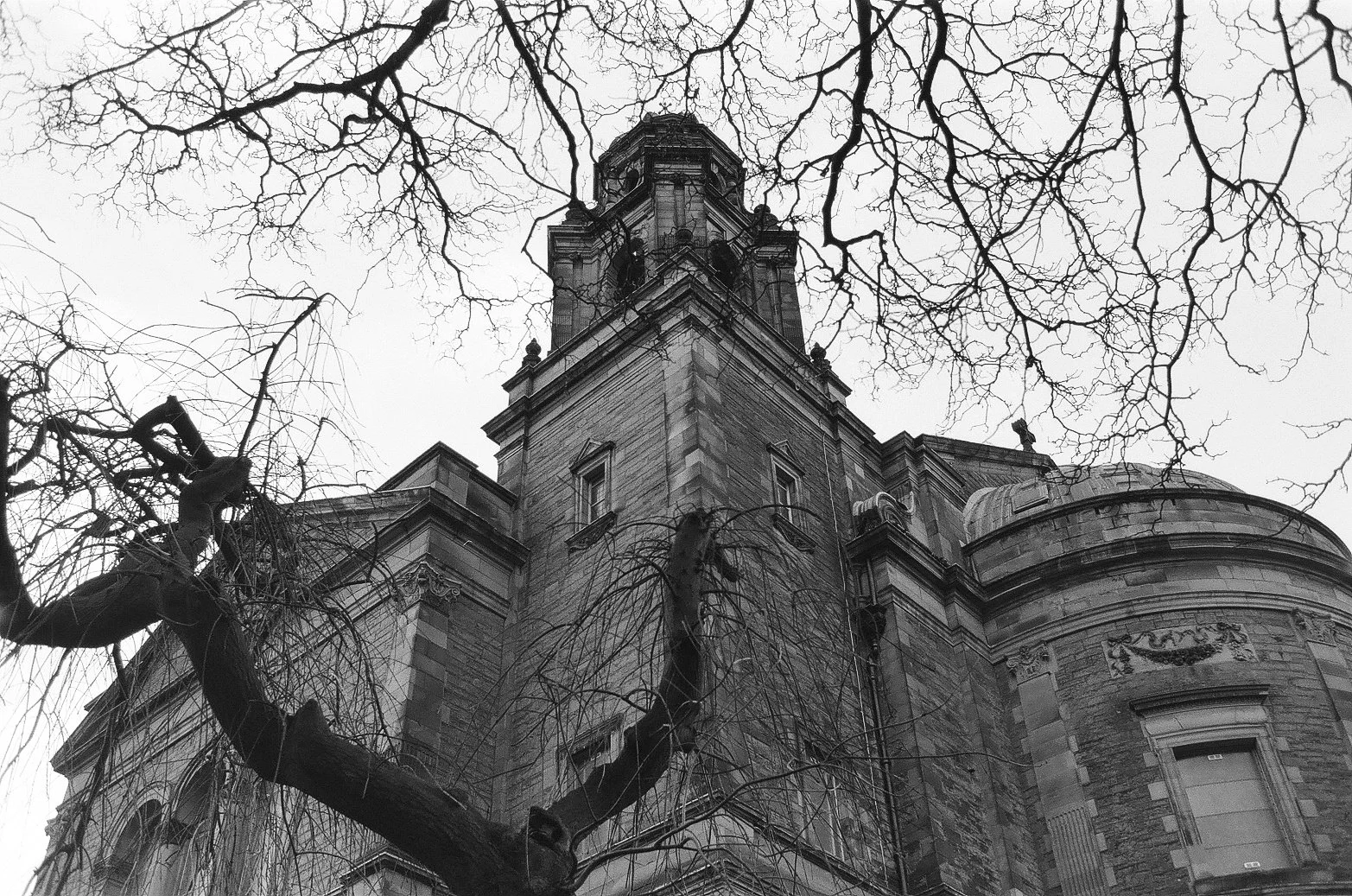 A black-and-white photo of a tall, old, stone building with a tower, viewed from below. Bare tree branches reach across the sky in the foreground.