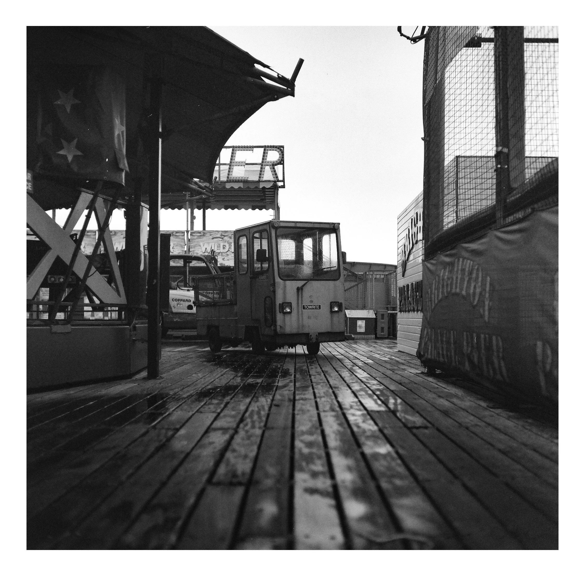 Black and white photo of an empty amusement park ride on wet wooden planks, featuring a small remote-controlled train in the center.