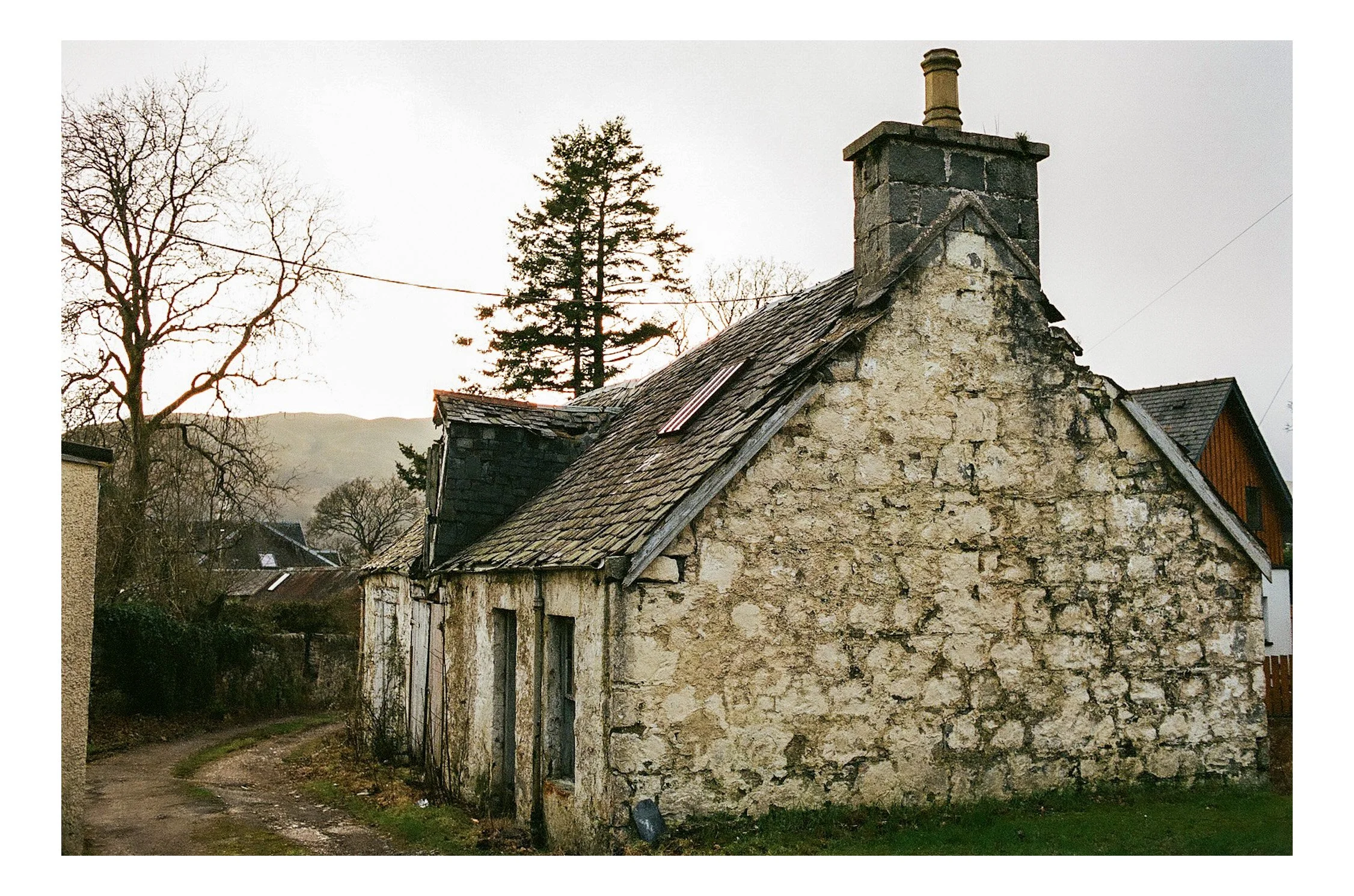 An old stone cottage with a weathered slate roof, a small chimney, and a dirt road in front, set in a rural landscape with leafless trees and rolling hills in the background.