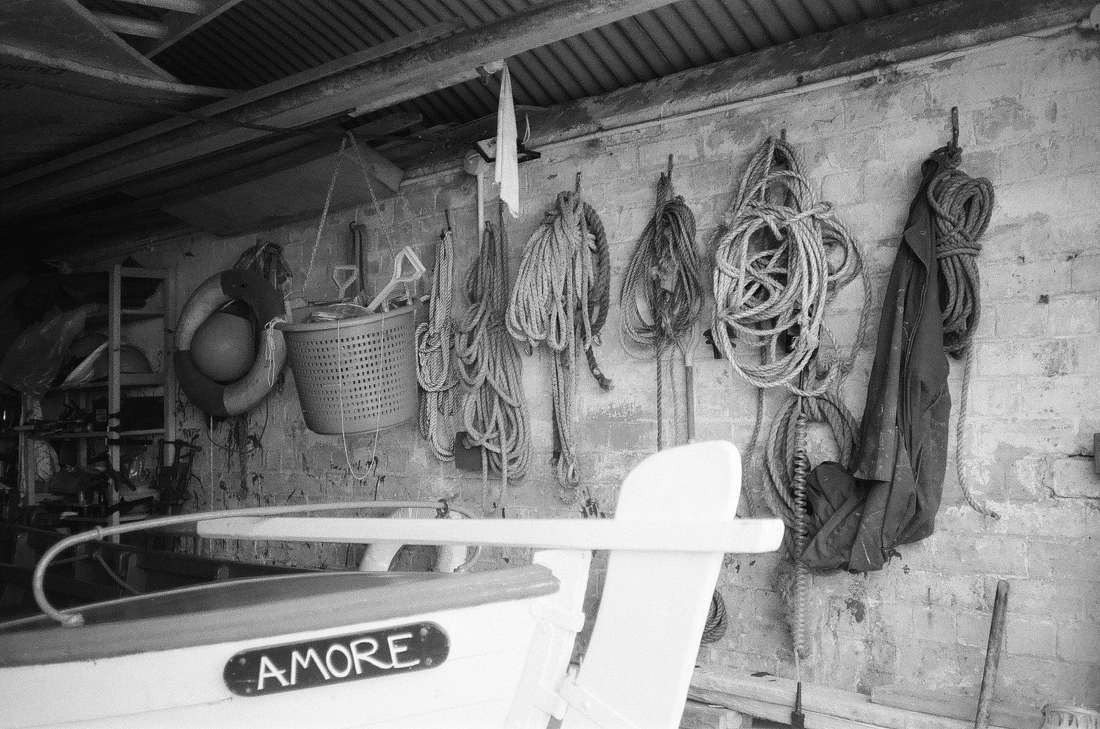 A black and white photo of a storage shed wall with nautical ropes, a life buoy, and various outdoor gear hanging. Part of a boat with the name 'A MORE' is visible in the foreground.