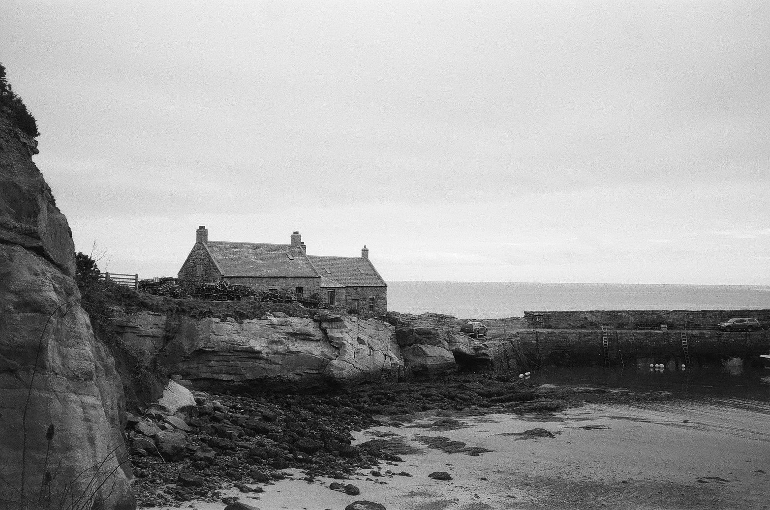 Black and white photo of a rocky coastline with a stone house on the cliff, a pier with cars, and the ocean in the background.