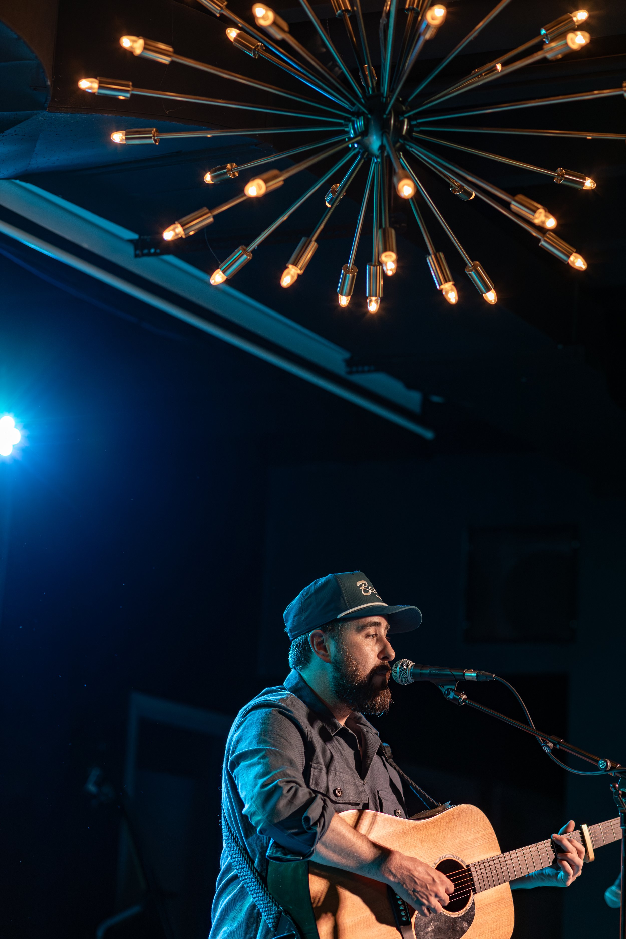A man with a beard wearing a dark cap and a dark button-up shirt is singing into a microphone while playing an acoustic guitar on stage, with a modern chandelier hanging from the ceiling above him.