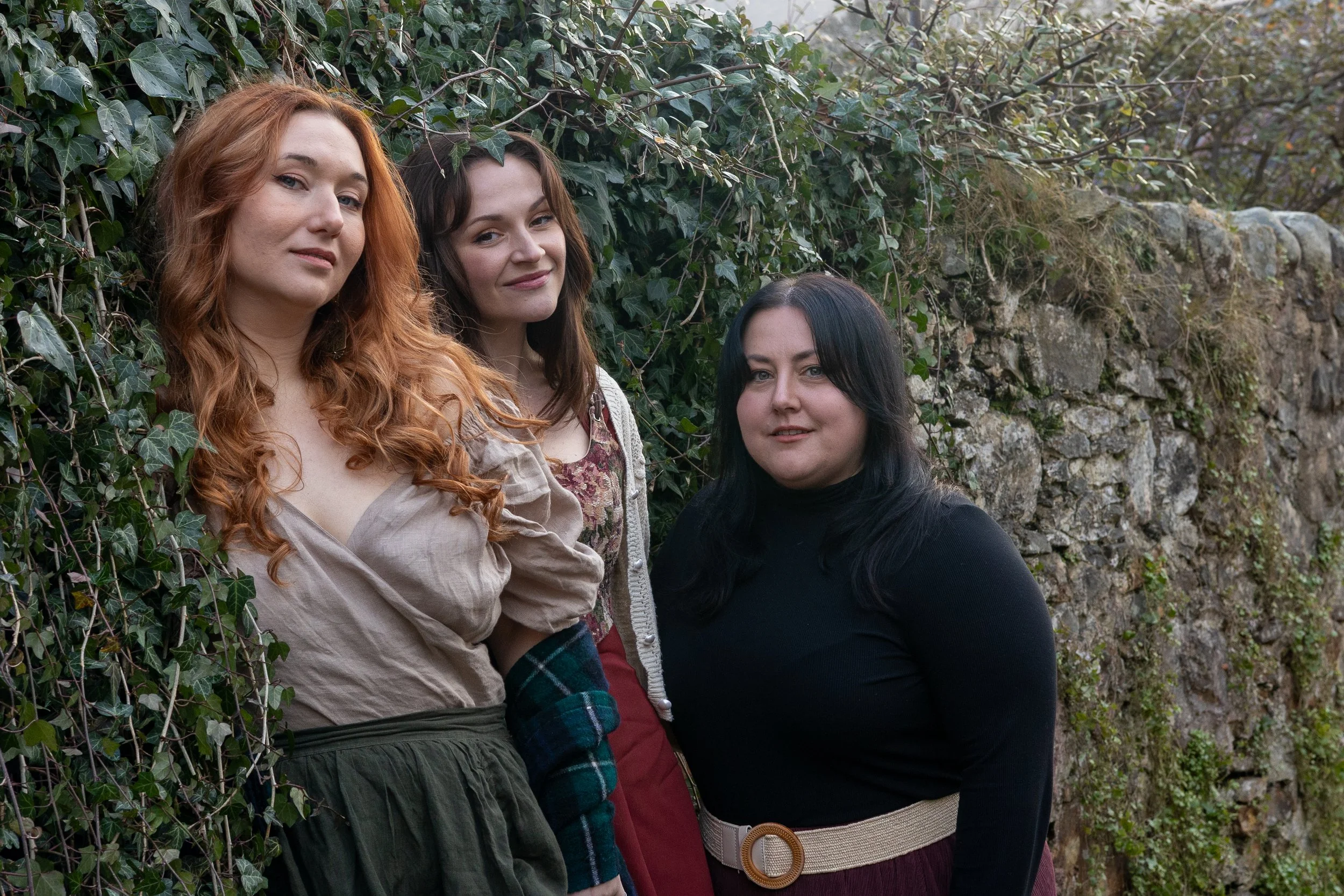 Three women standing outdoors near a stone wall covered in ivy, looking at the camera.