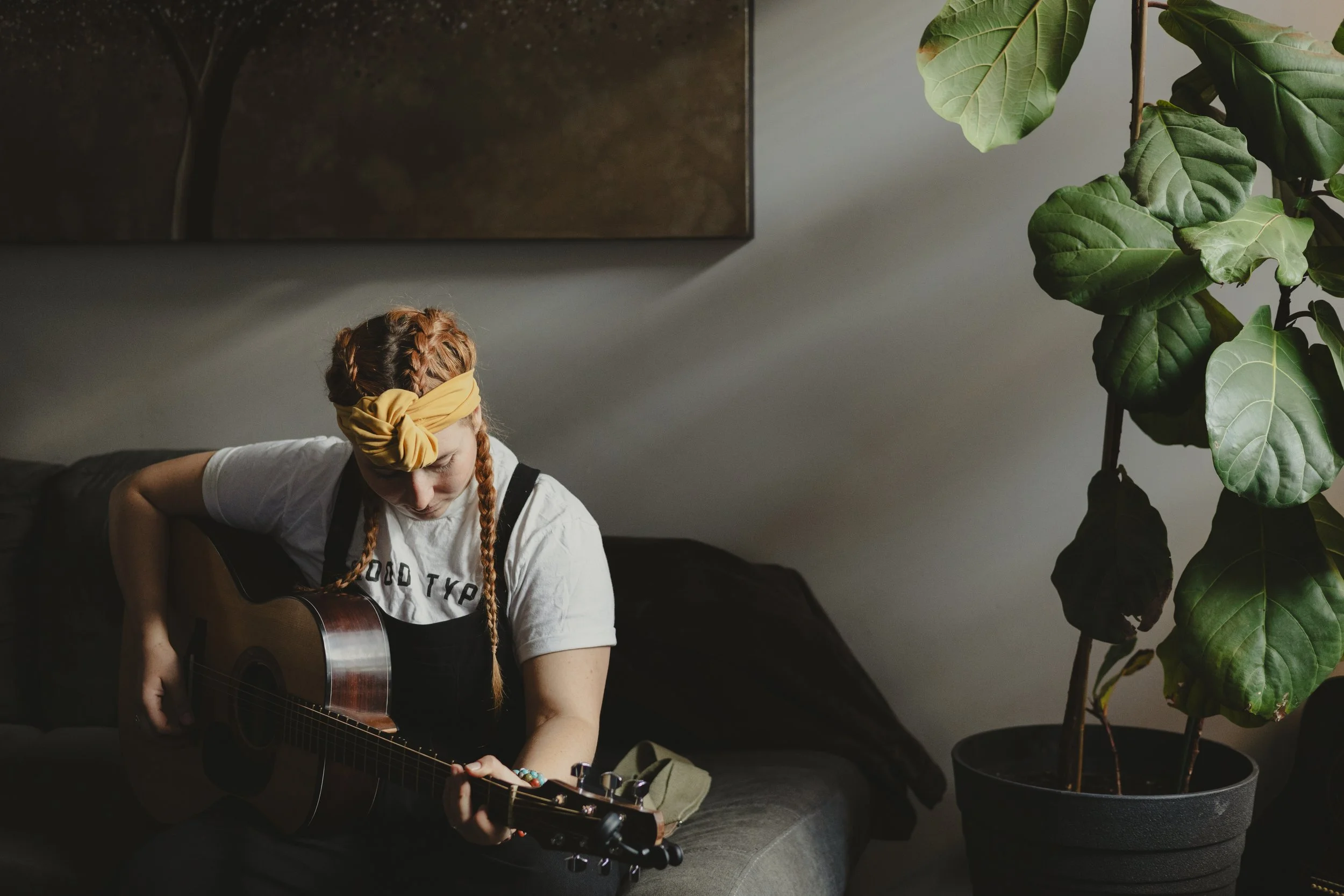 A young woman with red hair in braids, wearing a yellow head wrap and a white T-shirt, is sitting on a dark sofa playing an acoustic guitar. There is a large leafy plant in a pot and a dark pillow on the sofa.