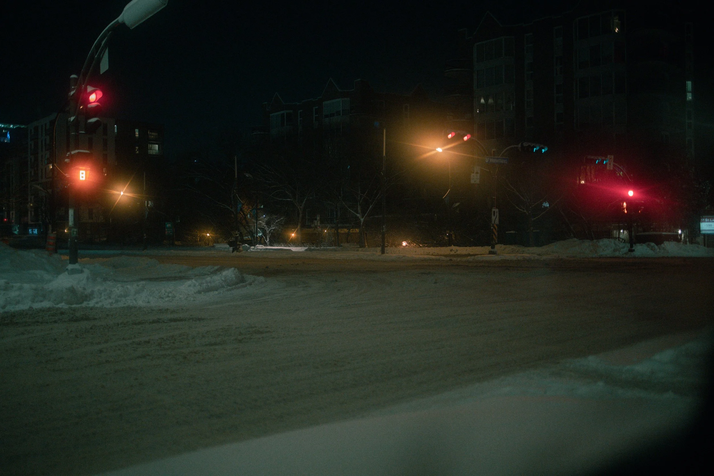 Nighttime city street view with snow-covered roads and buildings, traffic lights, and streetlights illuminating the scene.