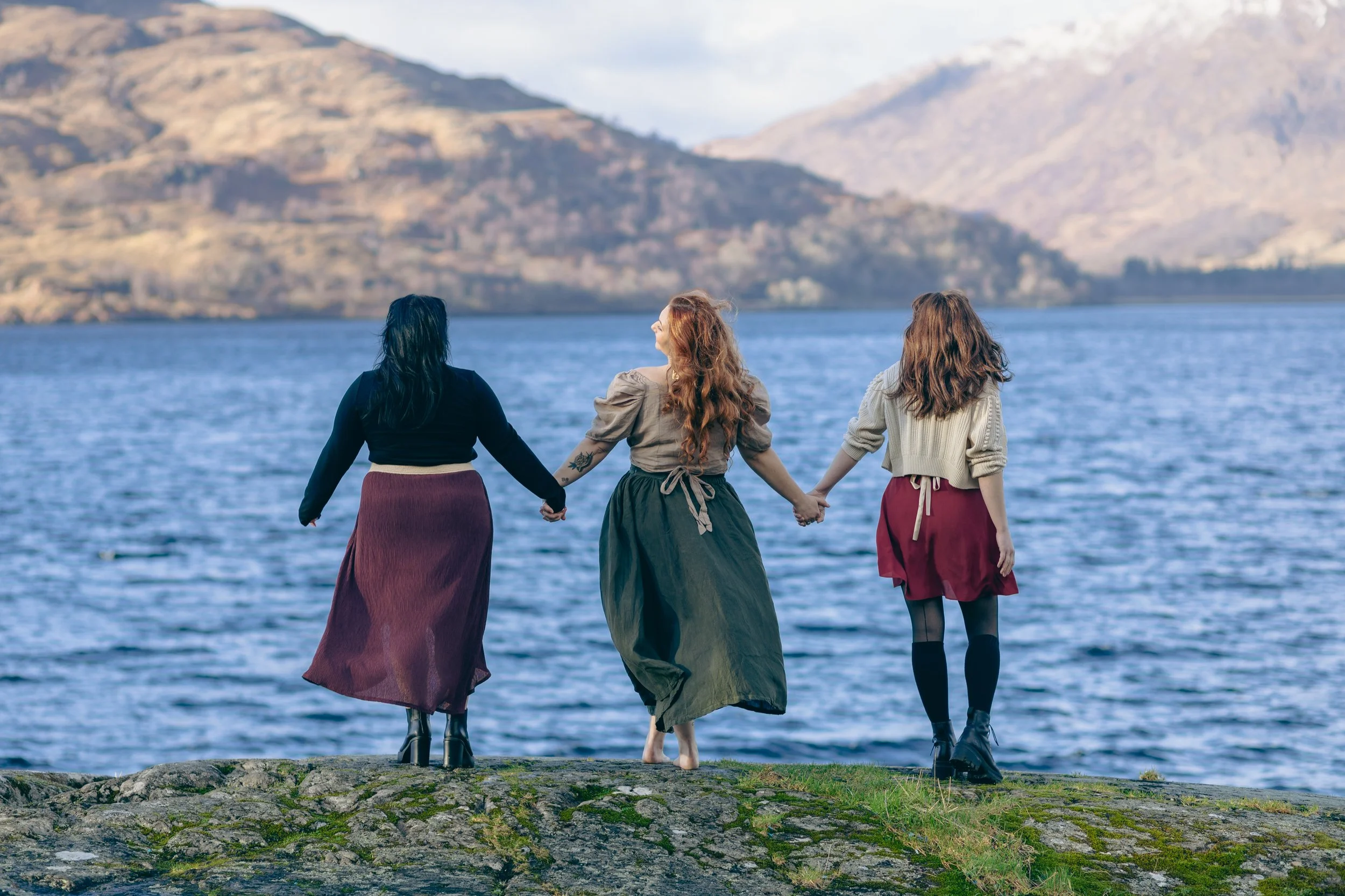Three women holding hands on a rocky shoreline by a lake with mountains in the background.