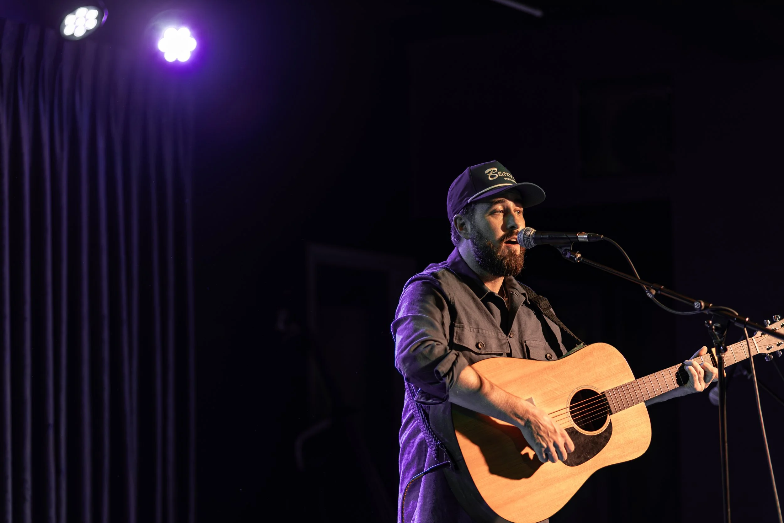 A man with a beard and wearing a black cap and dark shirt playing an acoustic guitar and singing into a microphone on stage with purple lighting.