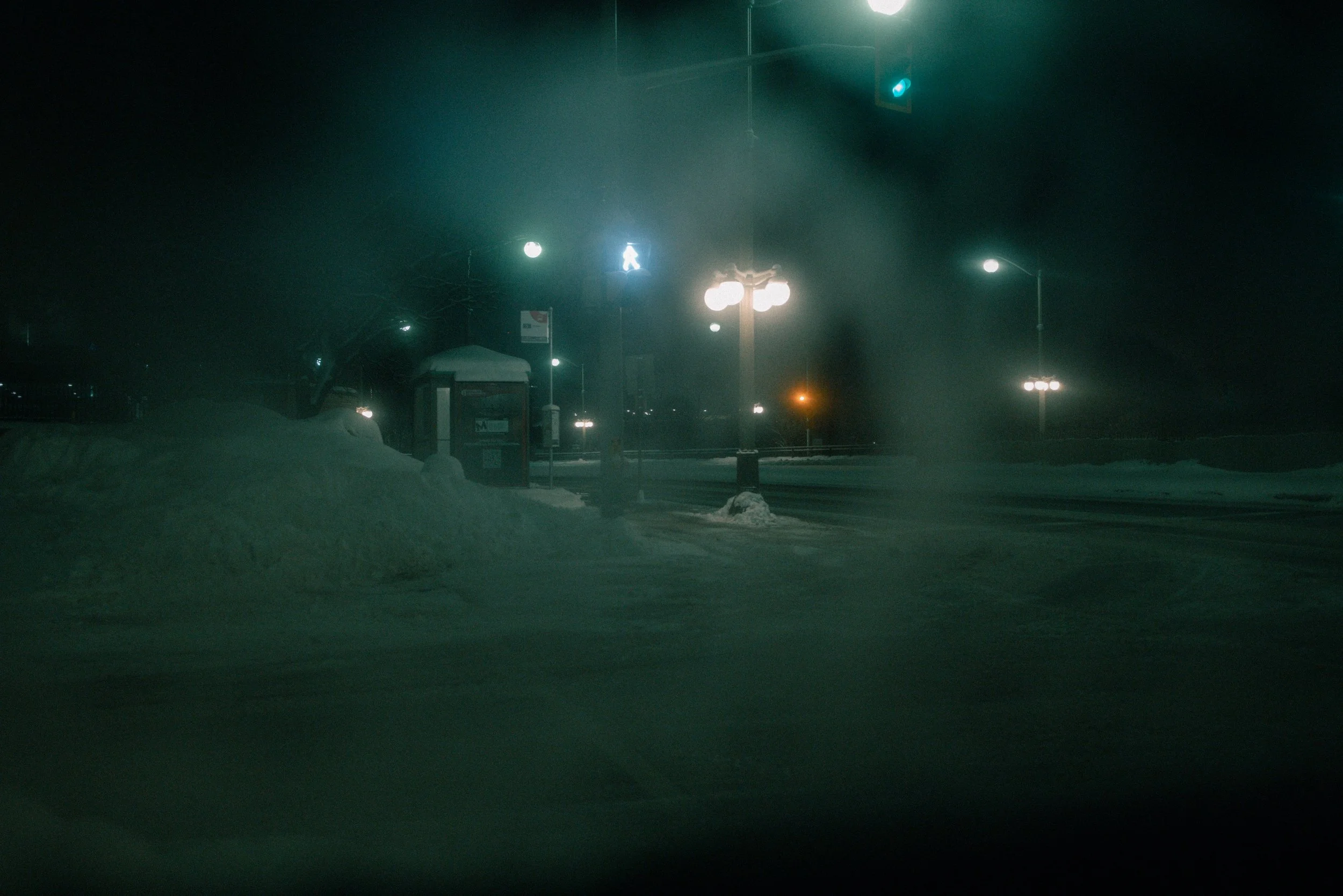 A snowy night scene of a street with green traffic lights, street lamps, a bus stop shelter, and snow-covered ground and piles of snow along the road.