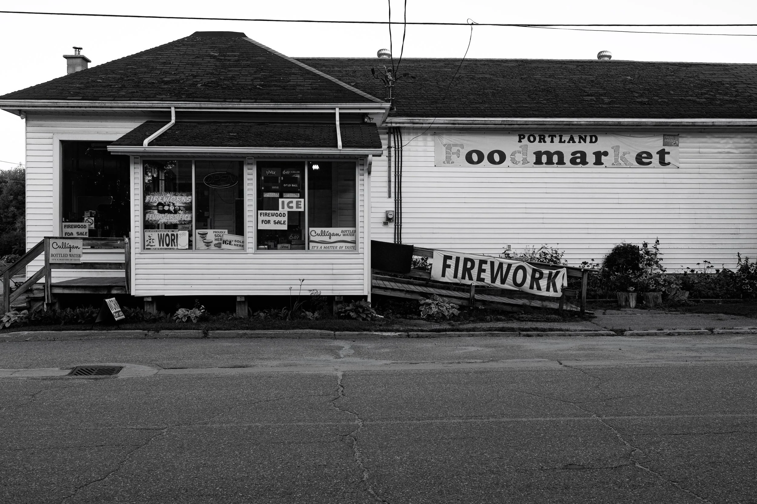 Black and white photo of a small storefront with a sign that reads 'Portland Food Market'. There are smaller signs in the window advertising firewood and bottled water for sale. The building has a sloped roof, and there is a sidewalk and street with 