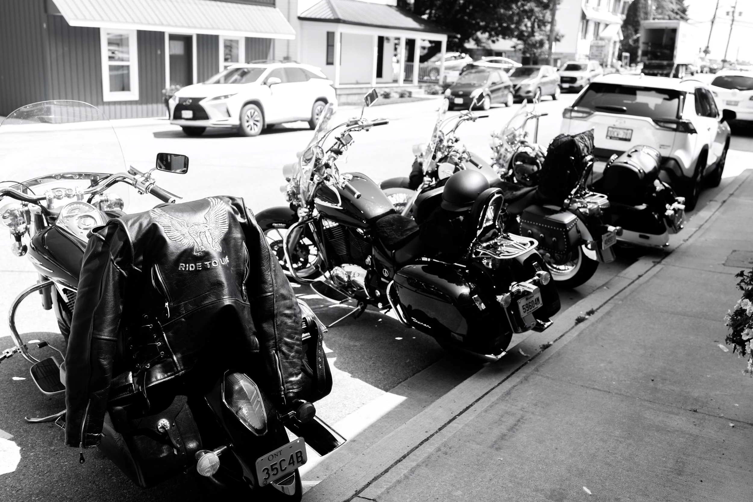 Black motorcycles with leather jackets and helmets parked along the sidewalk in front of houses and parked cars on the street in a sunny neighborhood.
