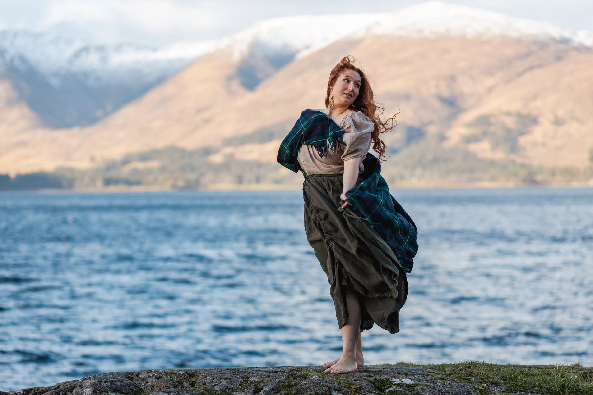 A woman with long, curly red hair stands barefoot on a rocky shoreline, holding her skirt while looking off to the side. She is wearing a beige blouse, a dark skirt, and a plaid shawl. Behind her, there is a large body of water with snow-capped mount