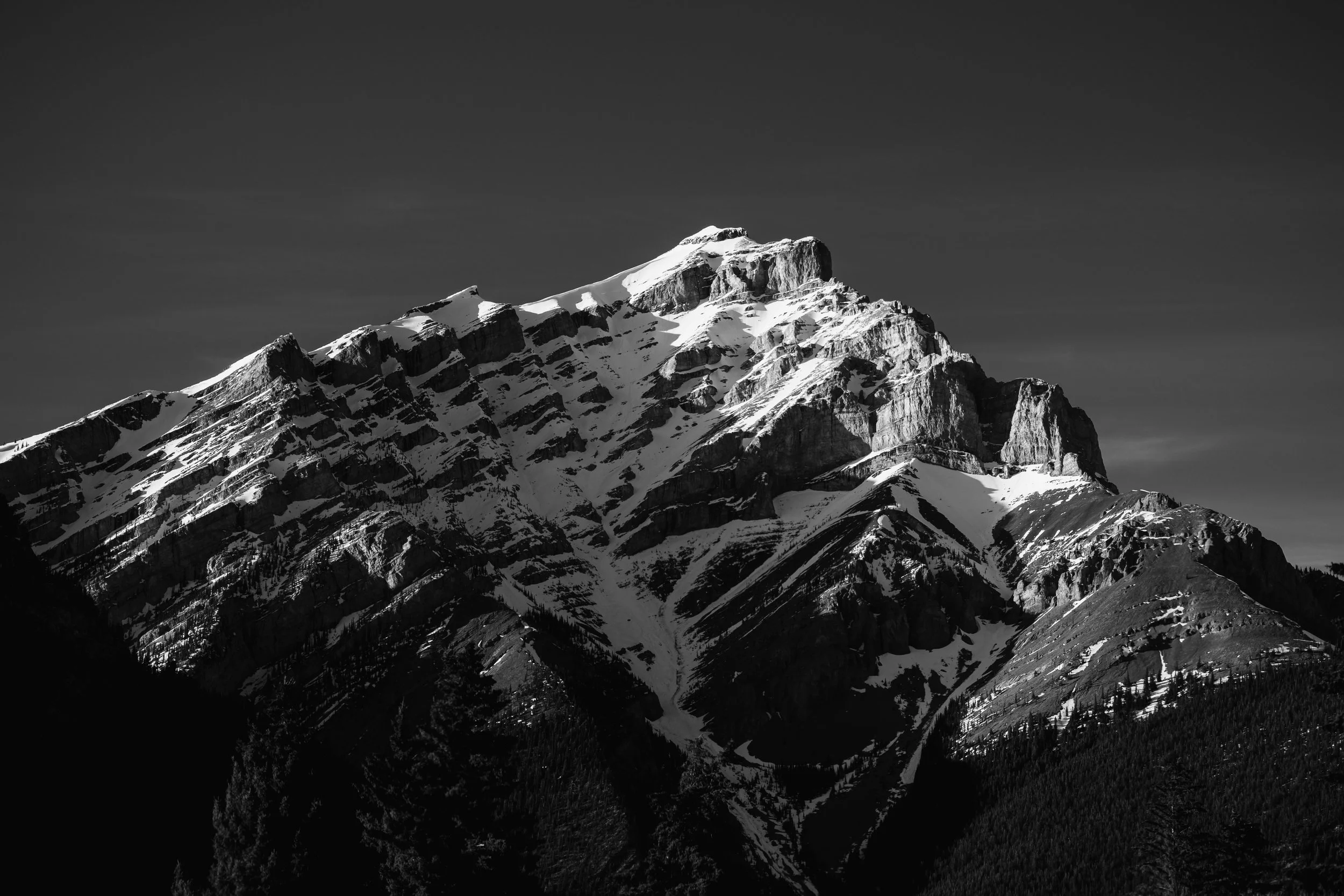 Black and white photograph of a snow-capped mountain with rugged terrain and a dark sky.