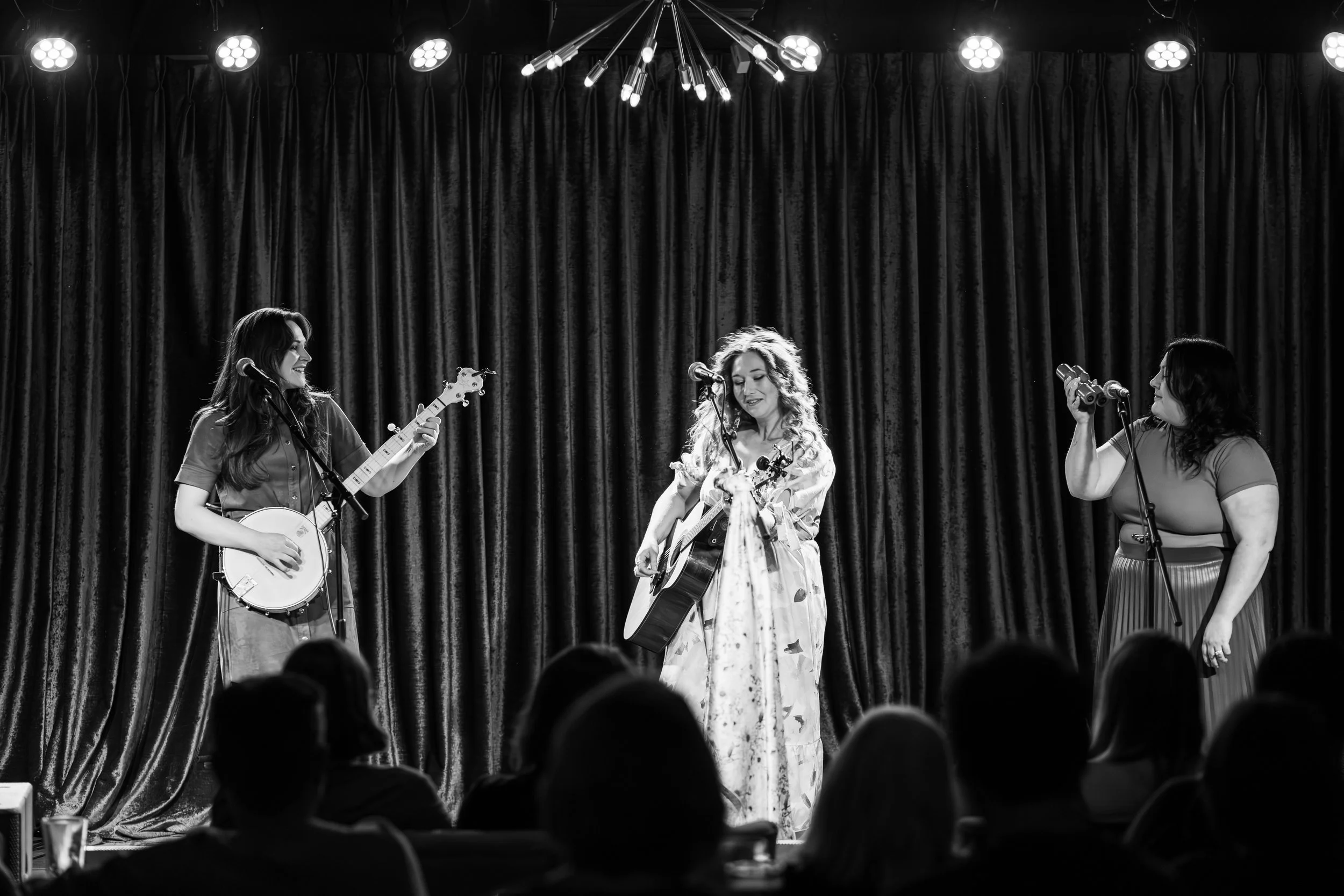 Three women performing on stage with microphones and musical instruments in front of a dark curtain, audience seated watching the performance.