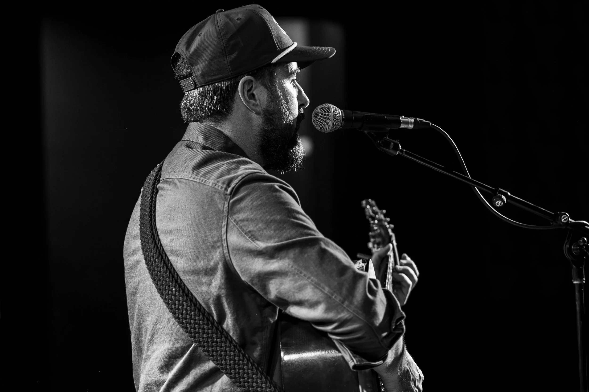 A black and white photo of a man with a beard, wearing a cap and a denim shirt, singing into a microphone and playing an acoustic guitar on stage.