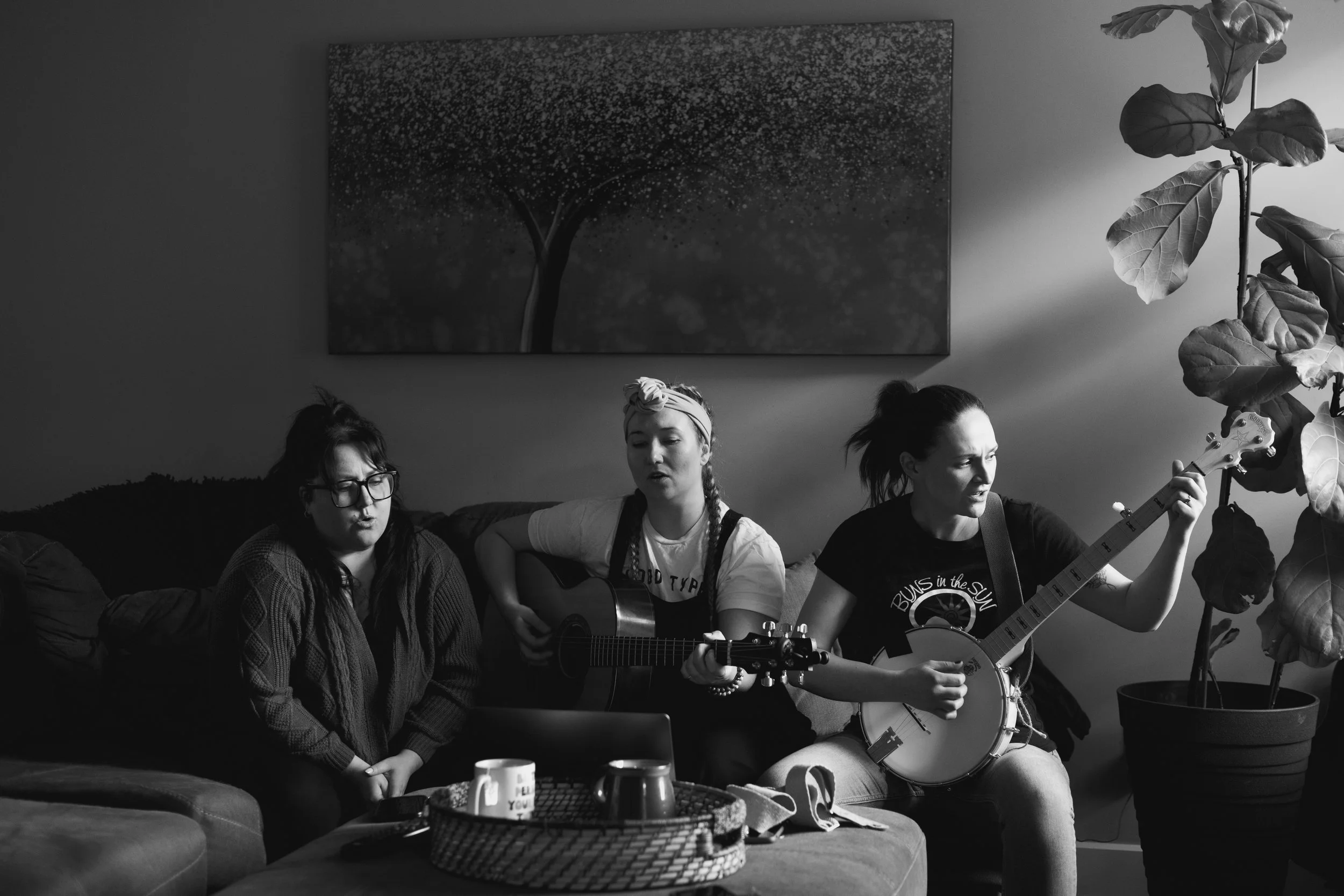 Three women sitting on a sofa, two of them playing guitars, in a living room with a large potted plant and a wall art painting.