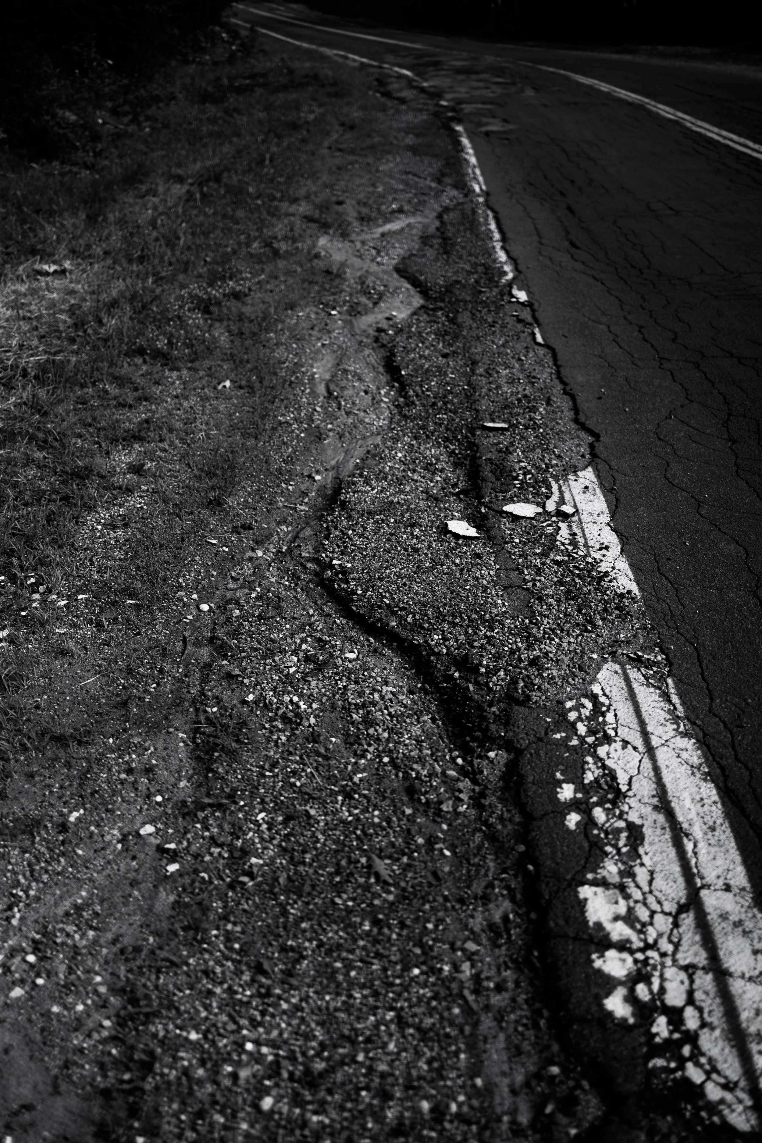 A crack in a cracked and damaged asphalt road alongside a rough dirt shoulder, at night.