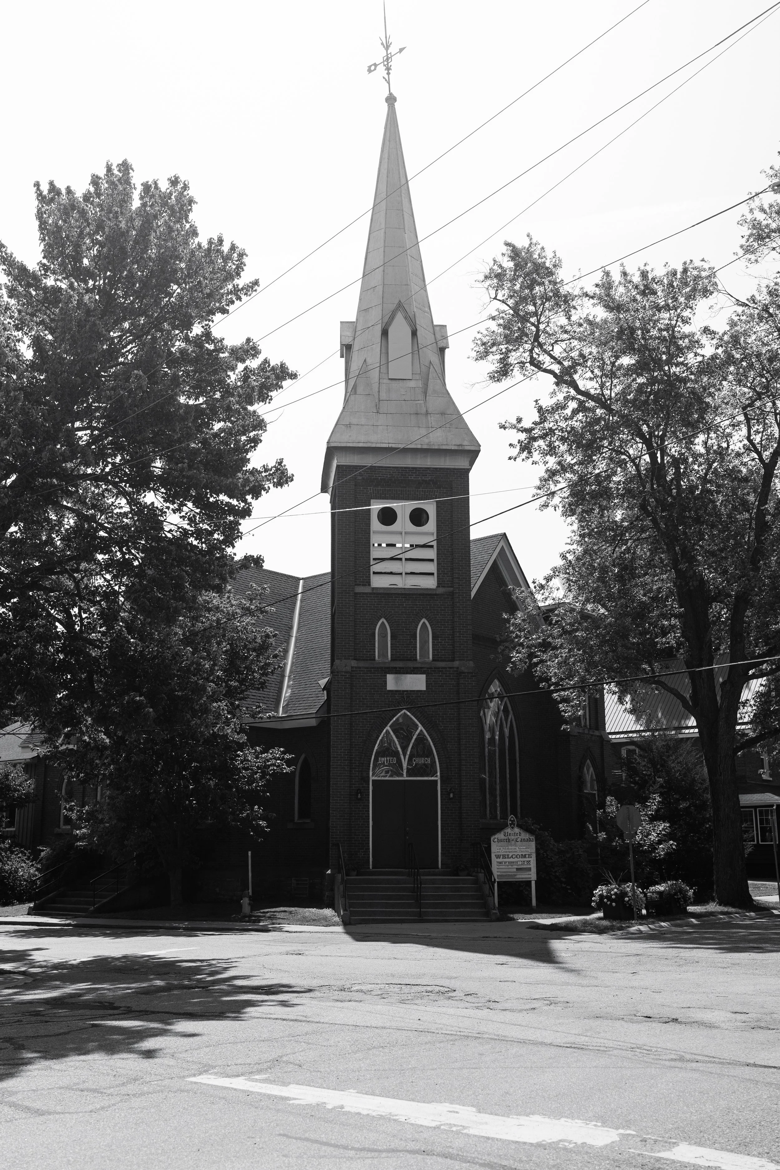 Black and white photo of a church with a tall steeple, trees surrounding it, and stairs leading to the entrance. The church has a sign that says 'United Church of Canada' and 'Welcome'.