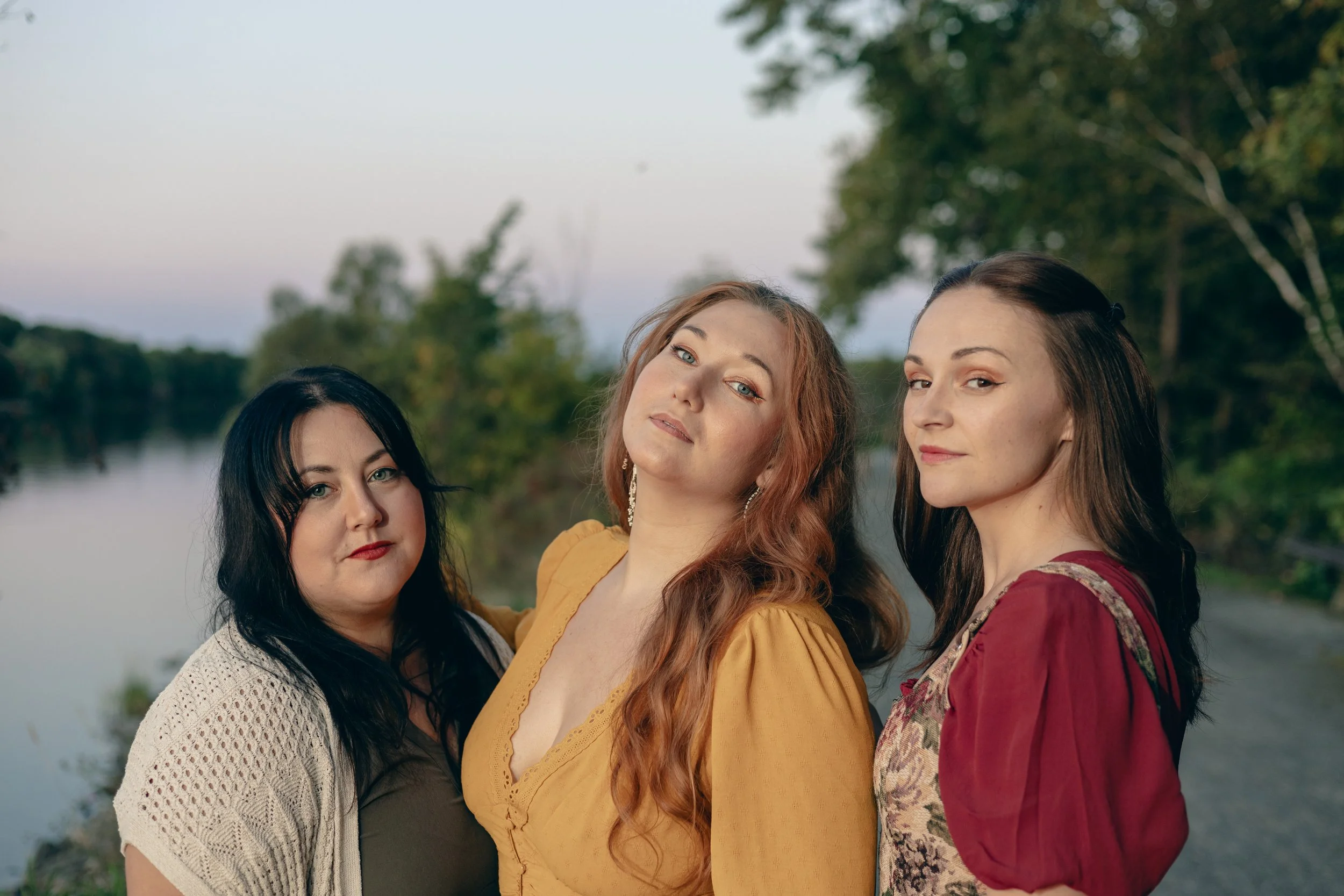 Three women standing outdoors near a river with trees in the background.