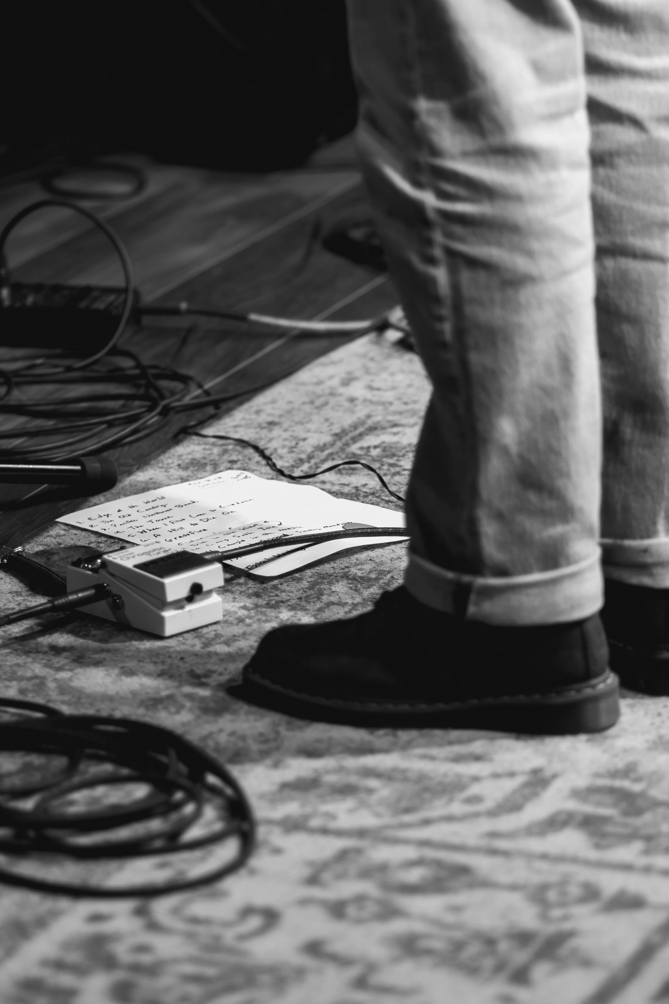 Close-up of a person standing on a carpeted floor, wearing jeans and black shoes, surrounded by microphone and audio equipment, with notes on a piece of paper on the floor.