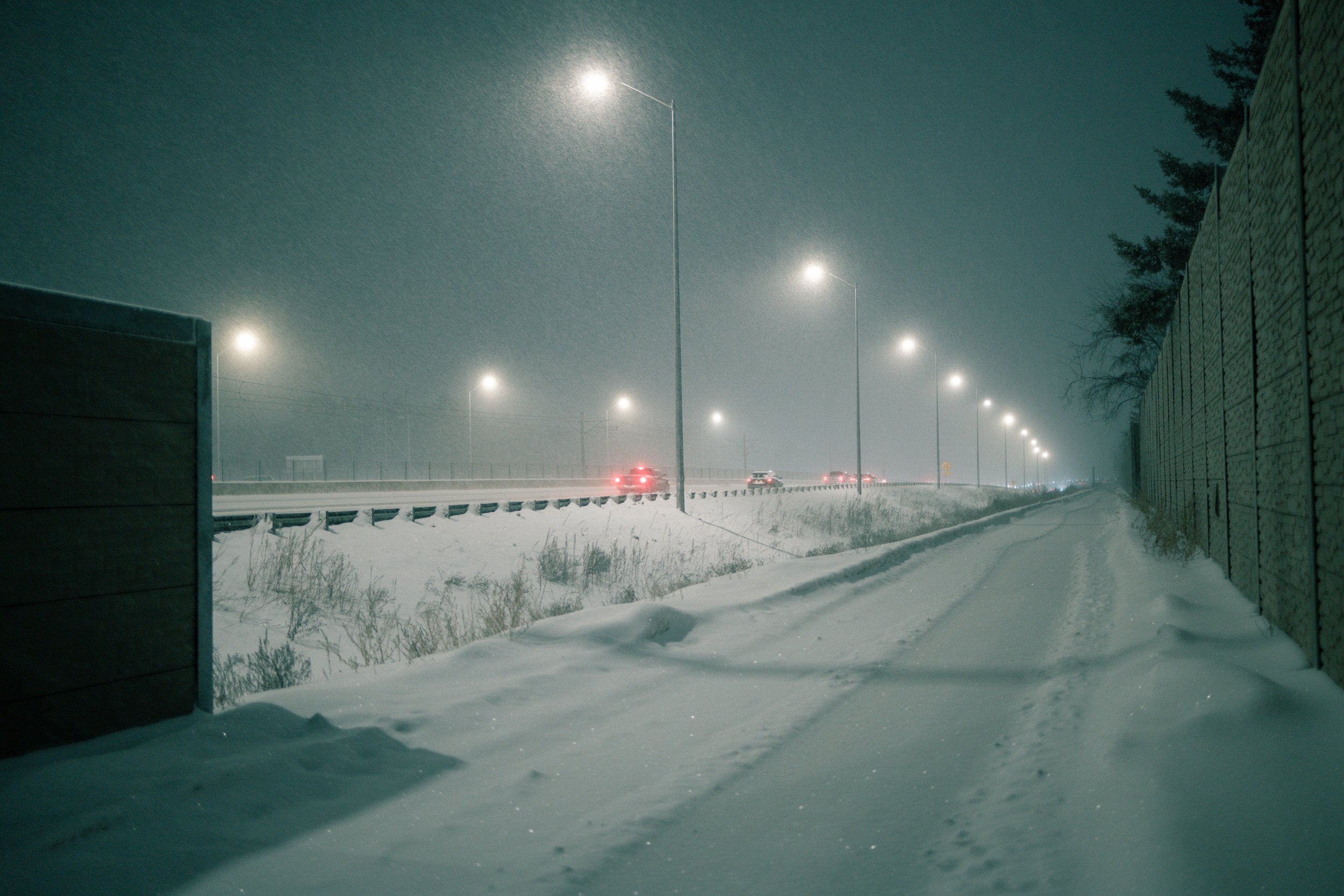 snow-covered road with cars and streetlights at night, snow falling, fence on the right side, dark sky.