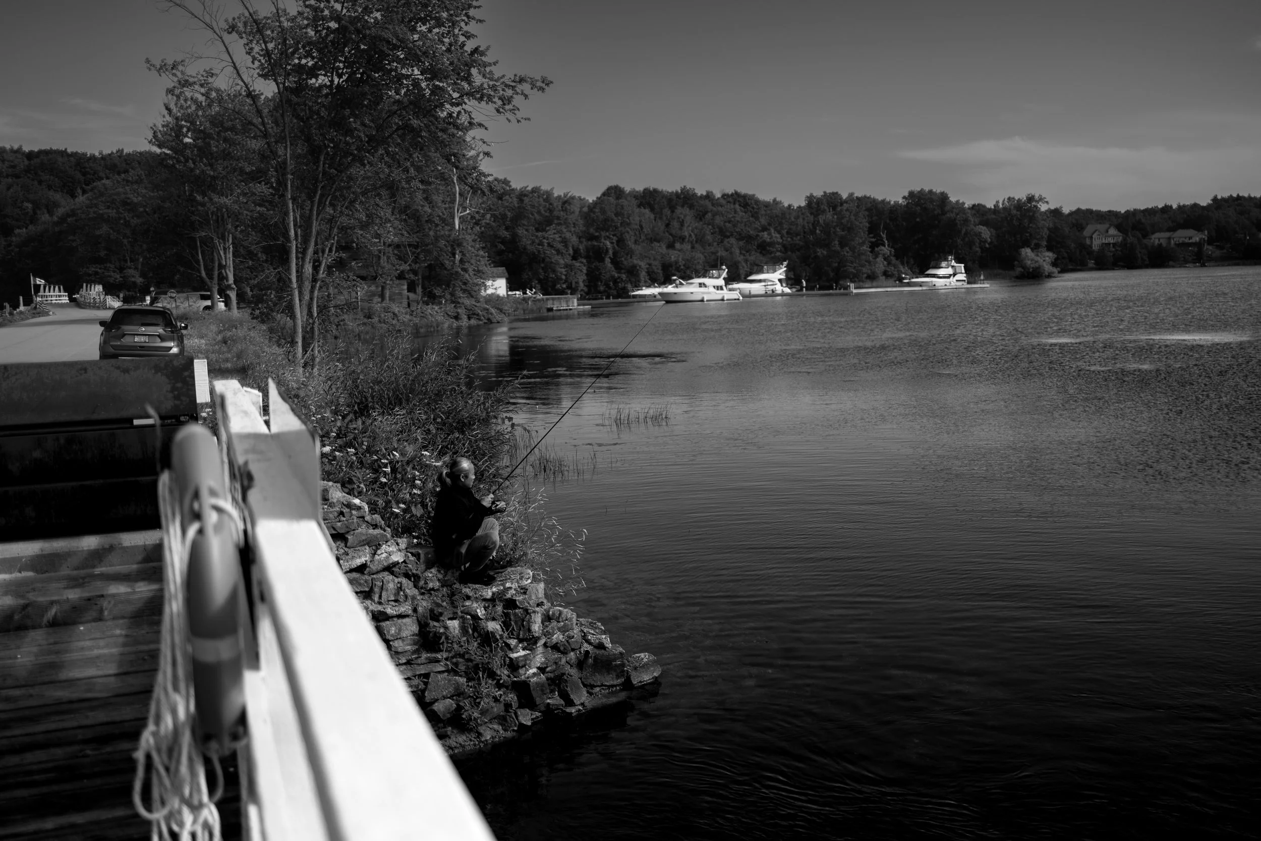 A person fishing by a lake with boats docked along the shoreline. Trees and houses are visible in the background, and the scene is in black and white.